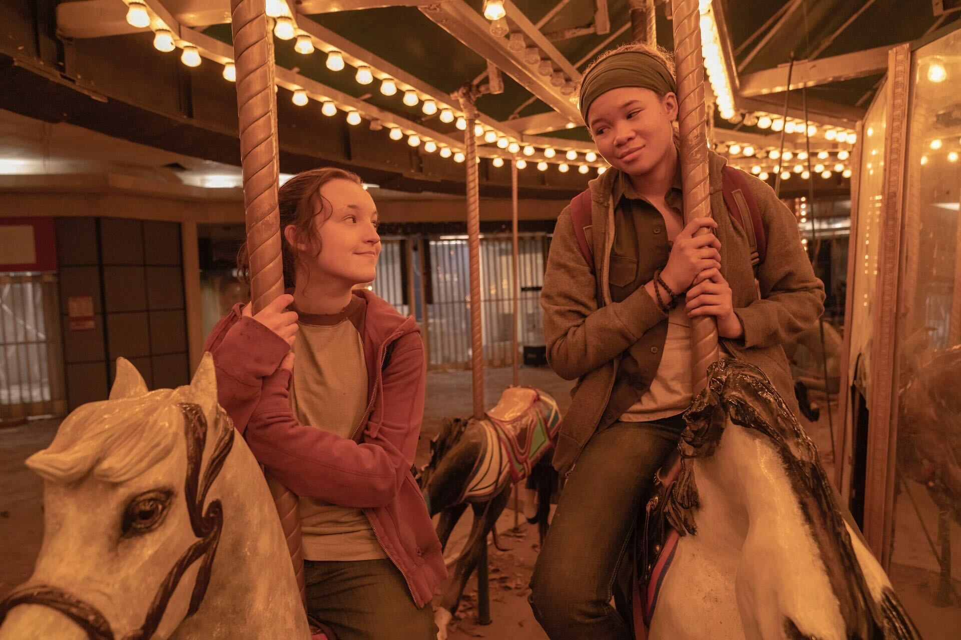 Bella Ramsey, left, and Storm Reid, right, look at each other with smiles as they ride fake carousel horses, bathed in the ride's warm light. 