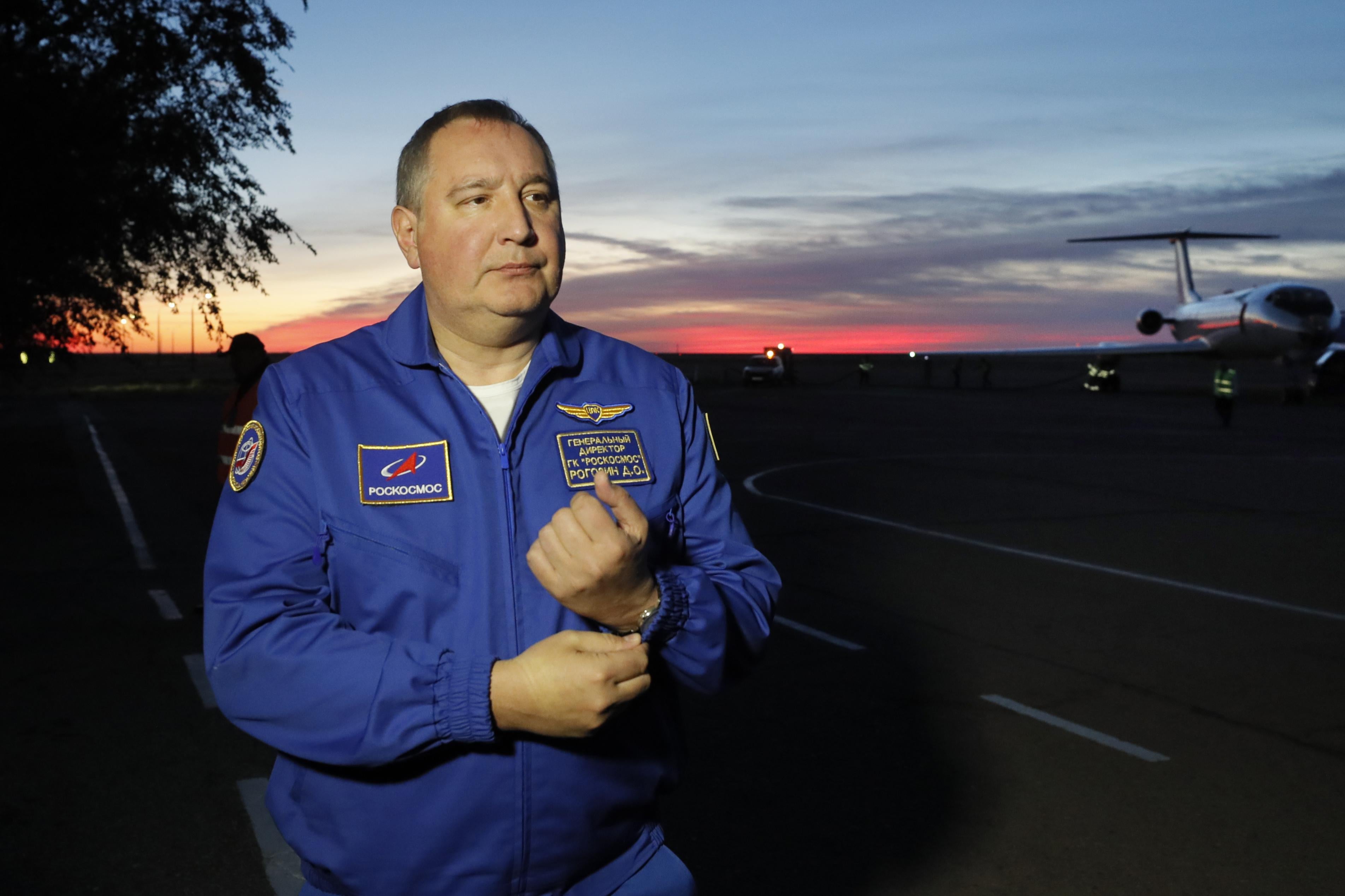 Dmitry Rogozin, the head of the Russian space agency Roscosmos, stands in front of a small jet plane while the sun sets behind him.