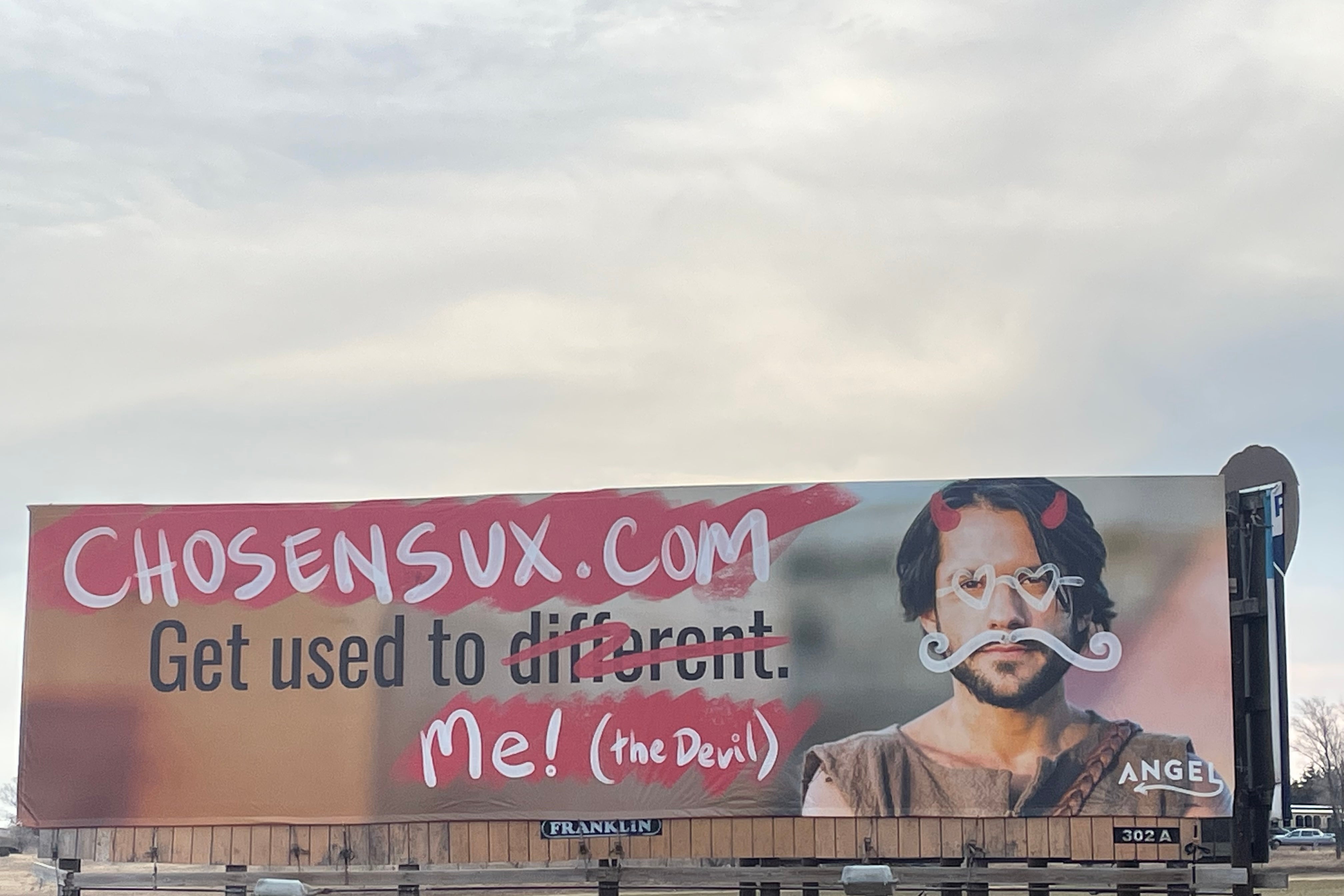 A billboard featuring the actor playing Jesus with graffiti of heart sunglasses, a twirly mustache, and devil horns on his face and the altered text "Get used to me! (the Devil)"