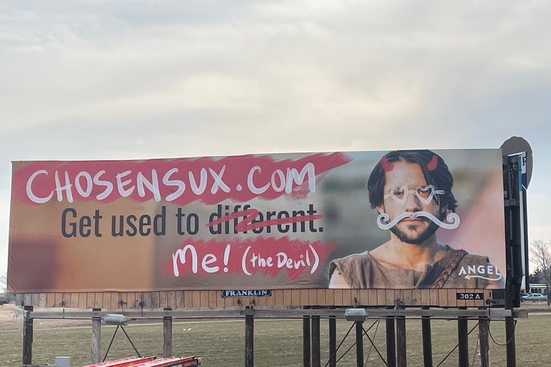 A billboard featuring the actor playing Jesus with graffiti of heart sunglasses, a twirly mustache, and devil horns on his face and the altered text "Get used to me! (the Devil)"