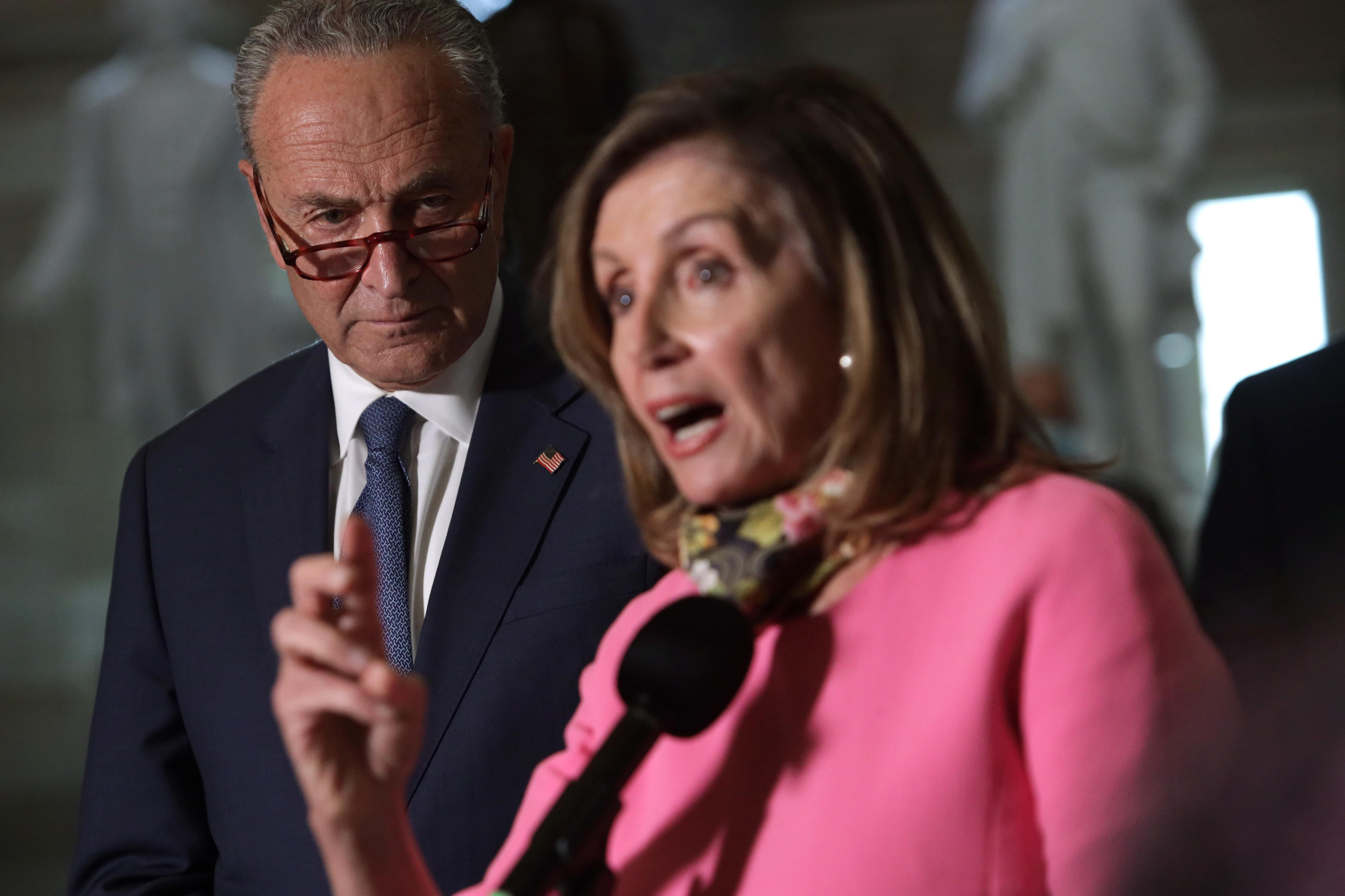 Schumer, glancing above his glasses, looks over Pelosi's shoulder as she speaks into a microphone.