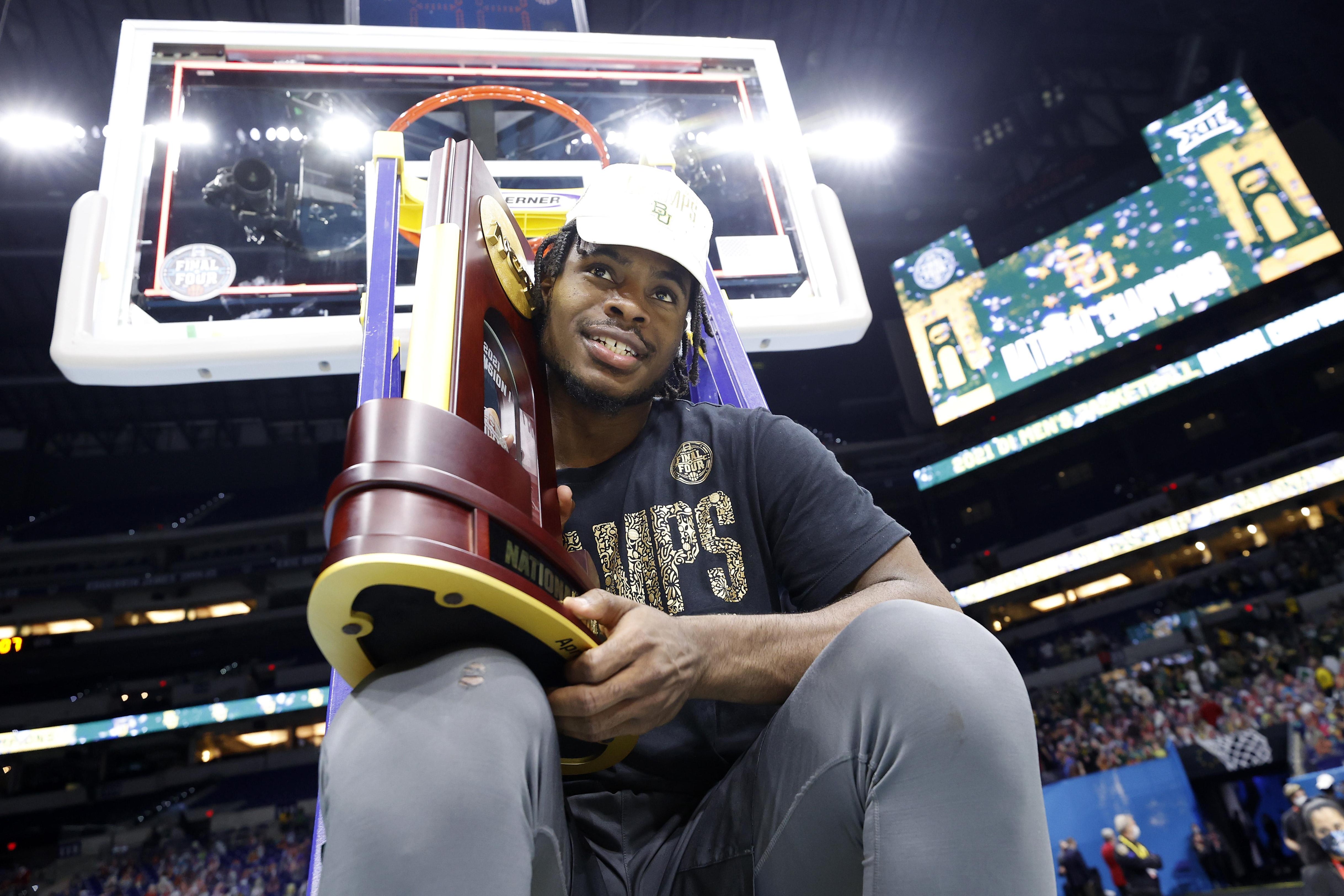 Baylor's Davion Mitchell smiling, sitting on a ladder under the hoop, with the NCAA title trophy