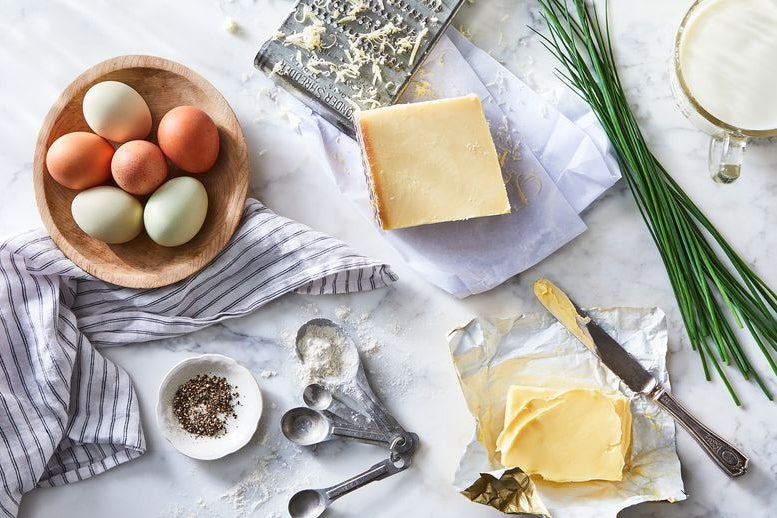 Ingredients laid out on marble countertop, clockwise from top left: bowl of six eggs, cheese grater and block of cheese, chives, butter, flour and pepper.