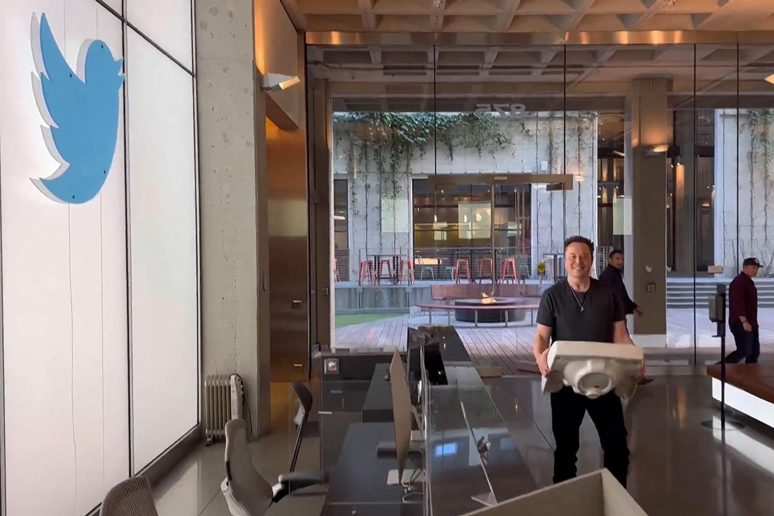 Elon Musk in an office with a Twitter logo on the wall; he is carrying a sink. 