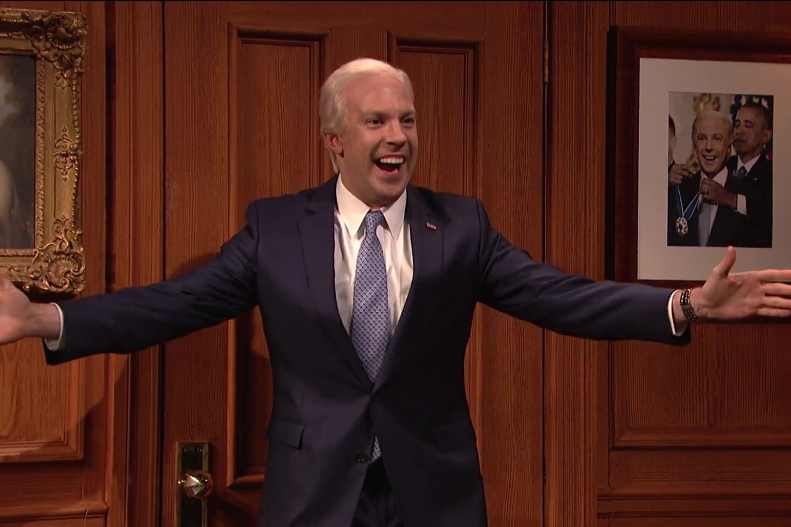 Jason Sudeikis, as Joe Biden, stands in front of a Photoshopped photo of Obama putting a medal around Sudeikis' neck as Sudeikis grins like a moron.