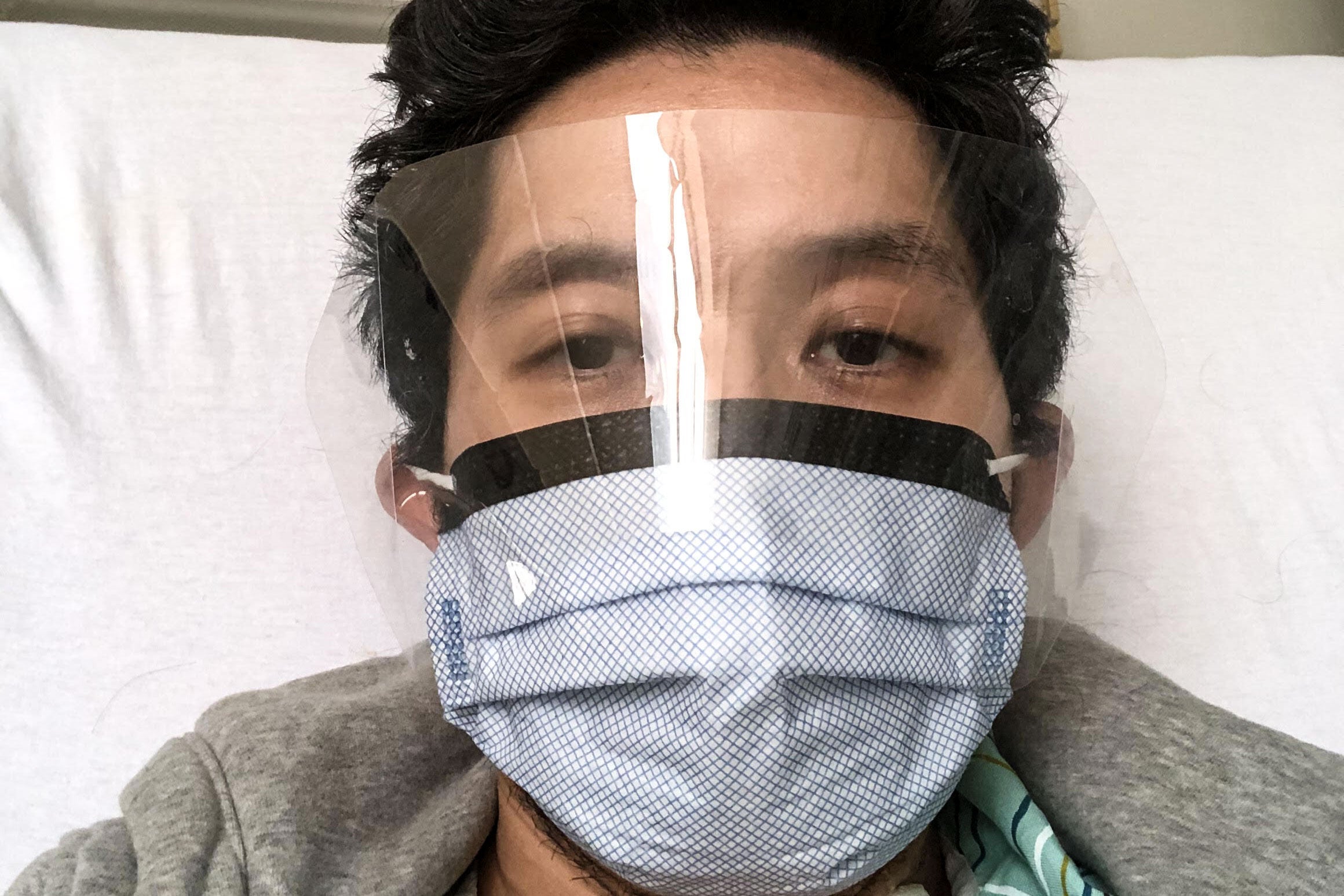 A man with his head on a white pillow wearing a face mask and face shield.