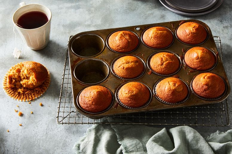 Muffins in a baking pan sit next to a cup of tea and a half-eaten muffin in its wrapper, crumbs spilling onto the countertop
