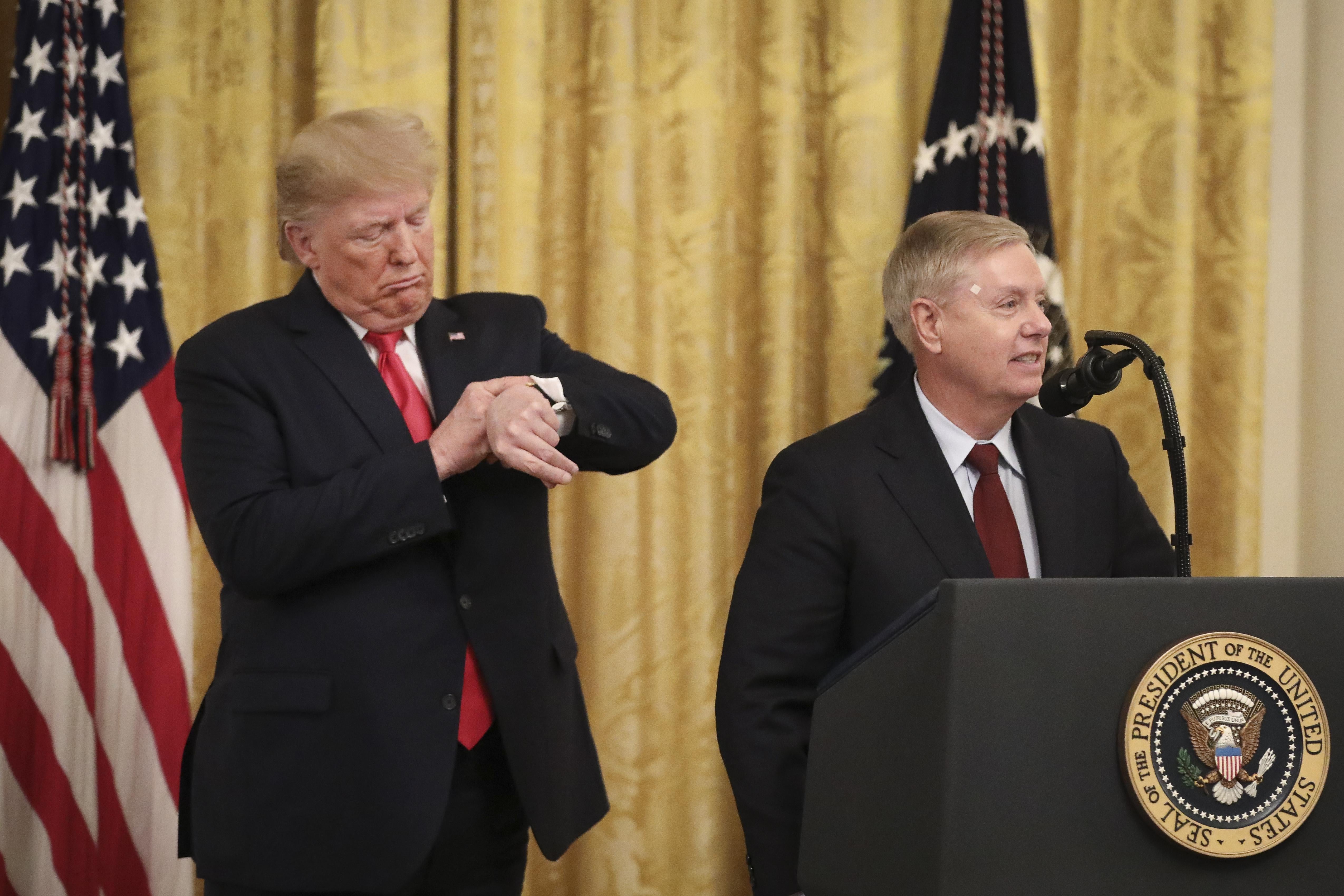 Donald Trump checks his watch  as he stands behind Lindsey Graham, who is at a lectern.
