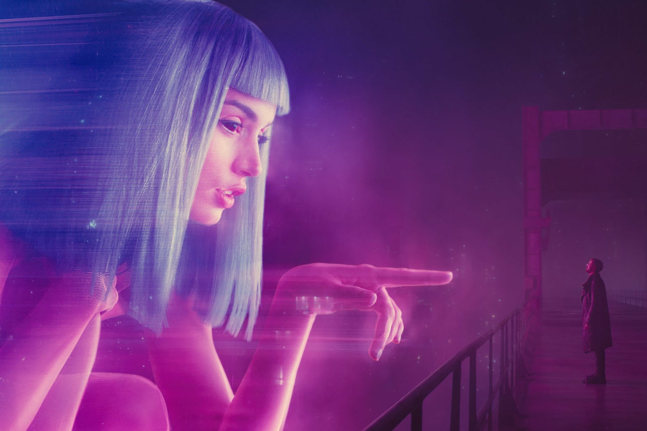 Ana de Armas is CGI modified into a large pink holograph of a woman with blue hair who points a large finger at human sized Ryan Gosling as he stands on a dark bridge in a dystopian society. 