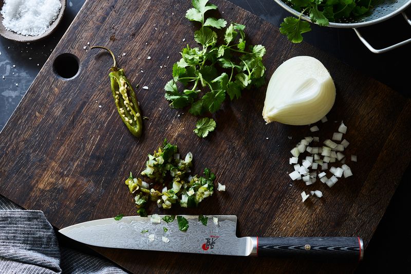 A knife sits next to a pile of minced herbs, onions, and peppers on a wooden cutting board.