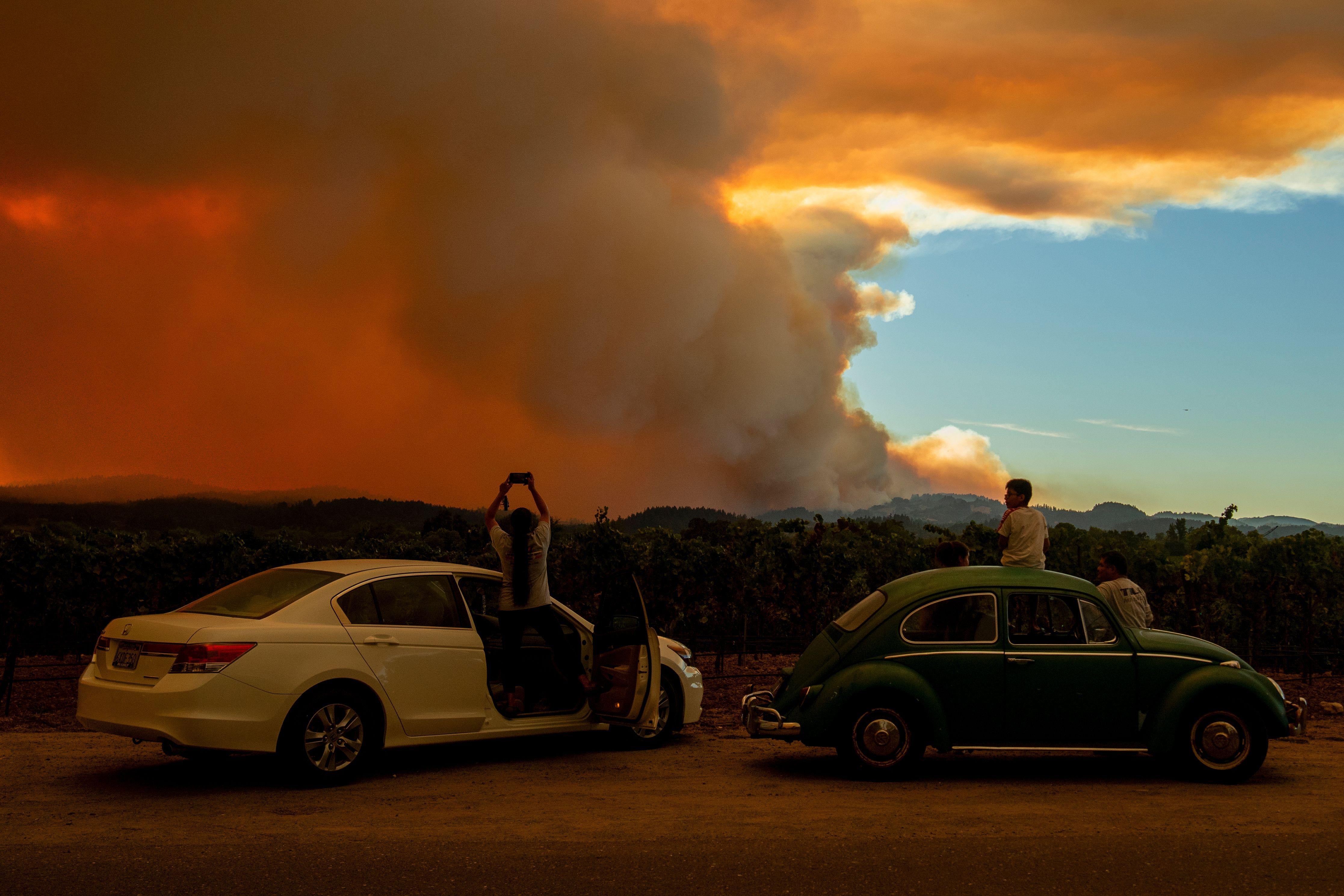People stand outside of two cars to watch as smoke takes over the sky.