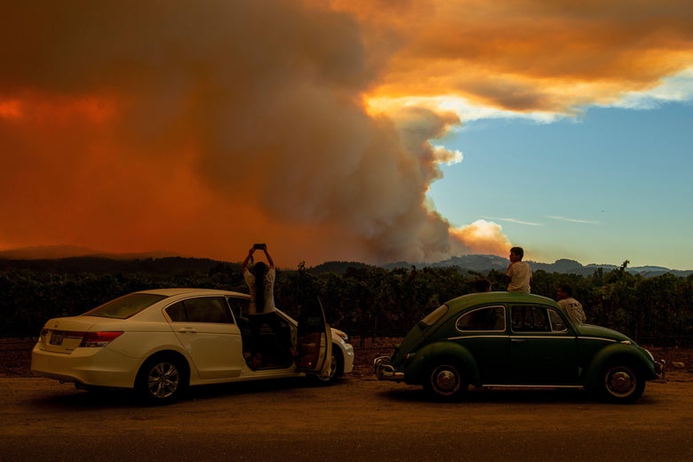 People stand outside of two cars to watch as smoke takes over the sky.