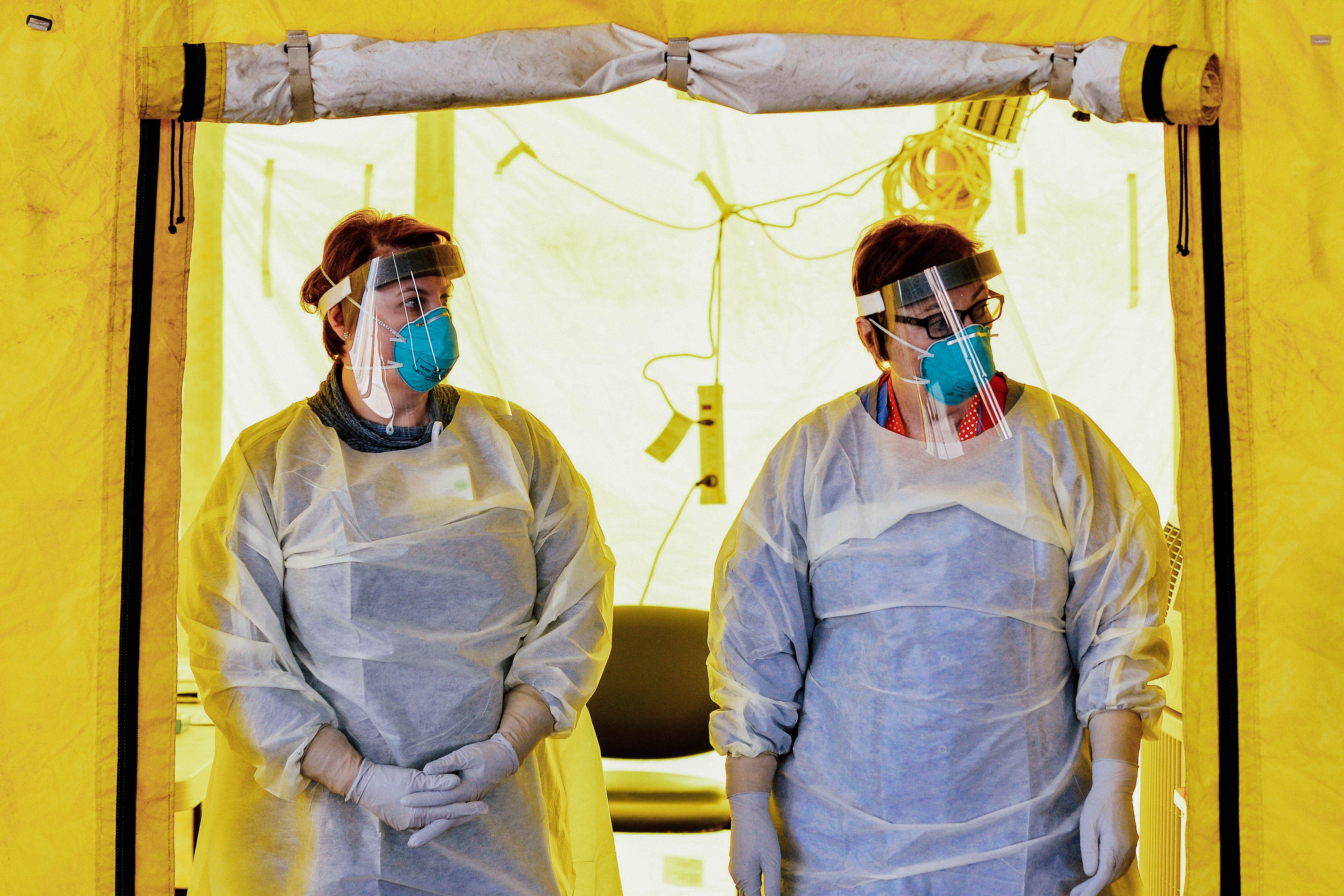 Two people wear masks and other protective gear in front of a tent doorway.
