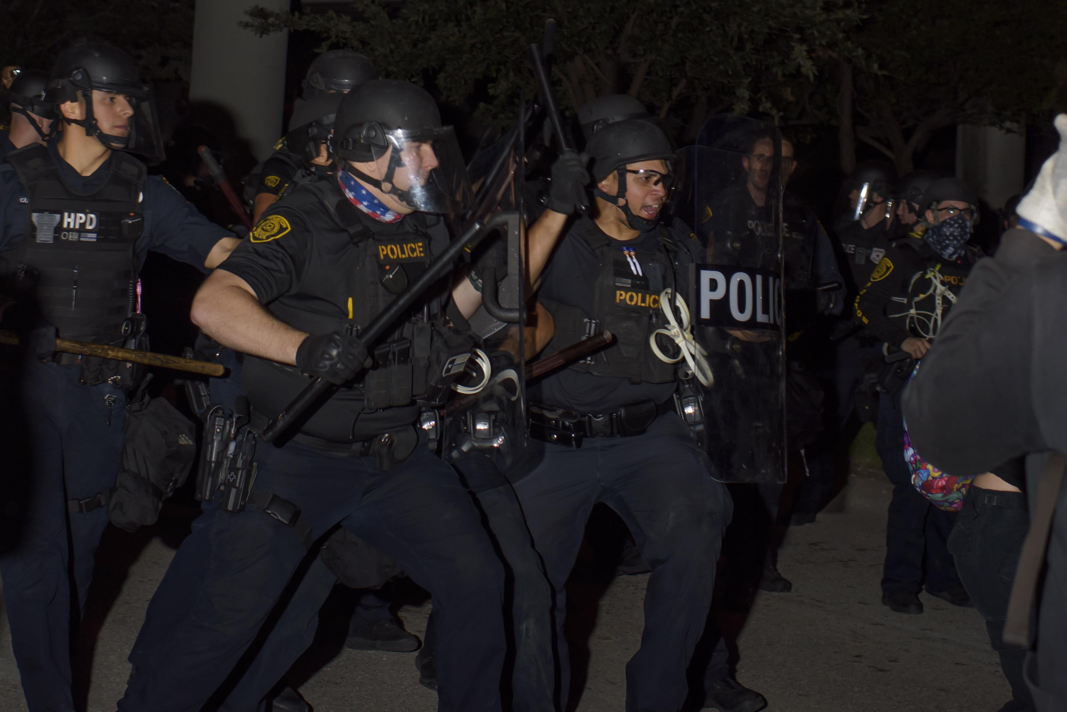 A group of police officers in riot gear with batons raised at night