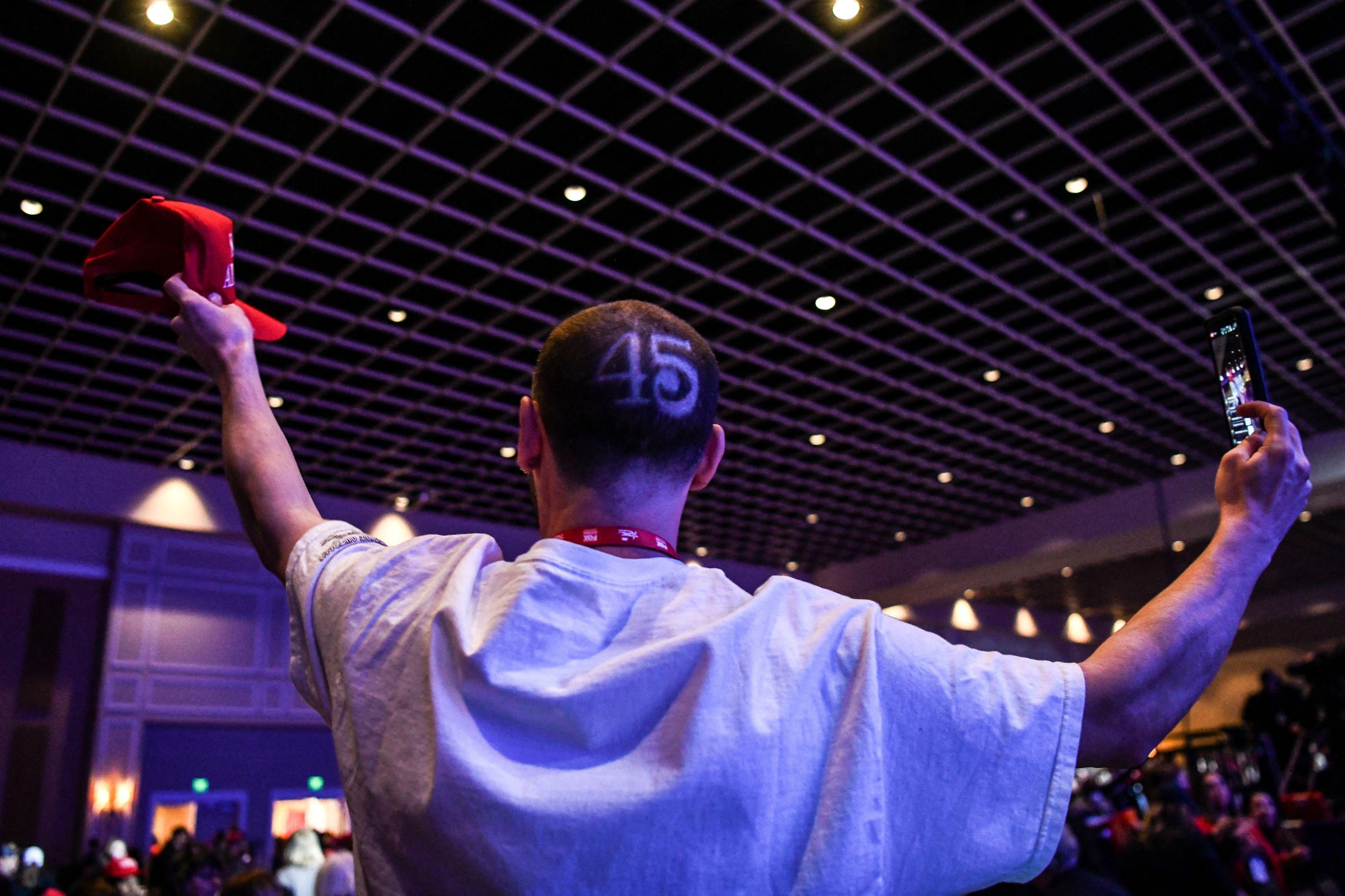 Man with the number 45 shaved onto his head shown from behind holding up a red MAGA hat in one hand and a phone in the other hand in a crowded room at CPAC