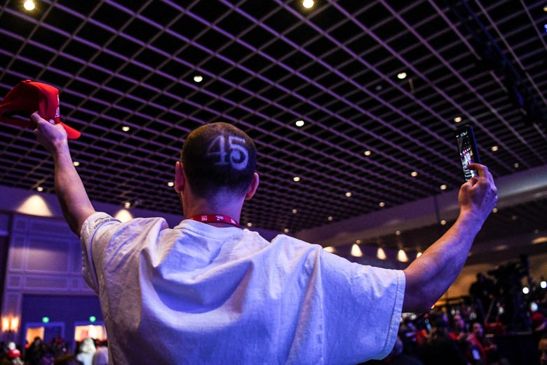 Man with the number 45 shaved onto his head shown from behind holding up a red MAGA hat in one hand and a phone in the other hand in a crowded room at CPAC