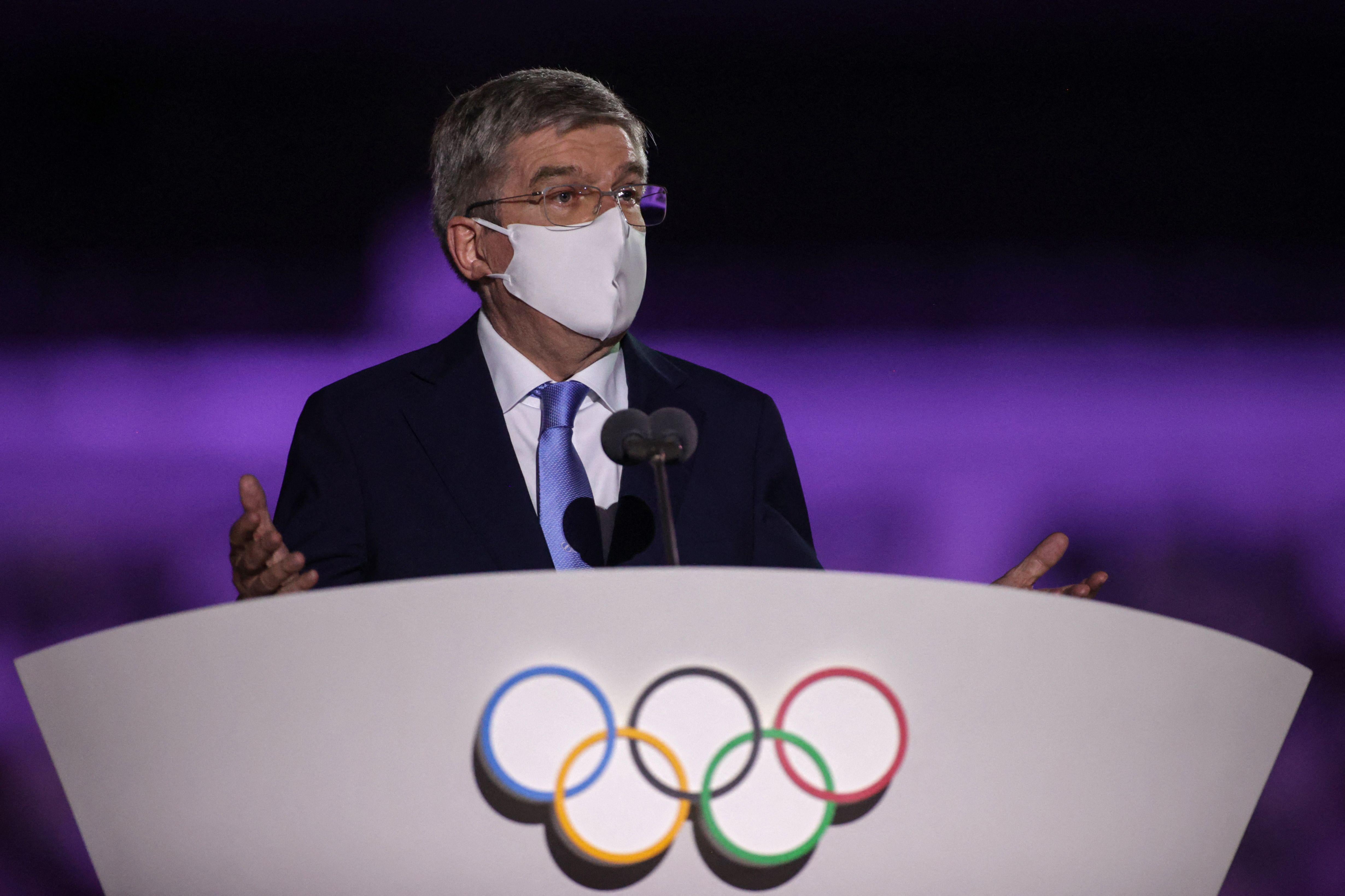 A masked older man in a suit and a mask speaks at a podium with the Olympic rings on it, gesturing with both hands