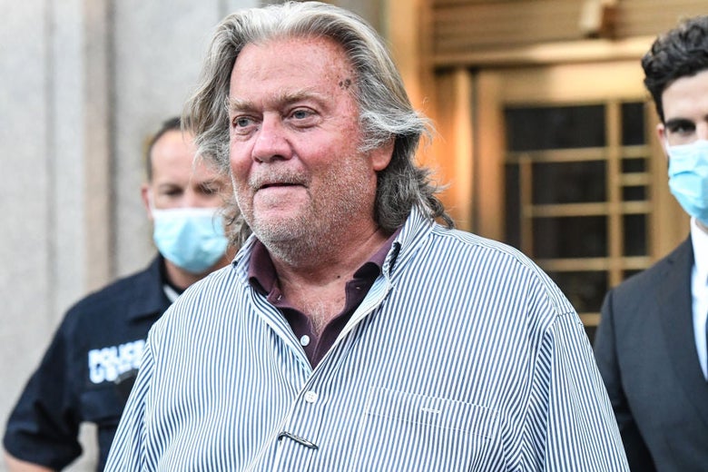 A long-haired Bannon, trailed by a law enforcement officer and a man in a suit, walks away from a door to the courthouse.