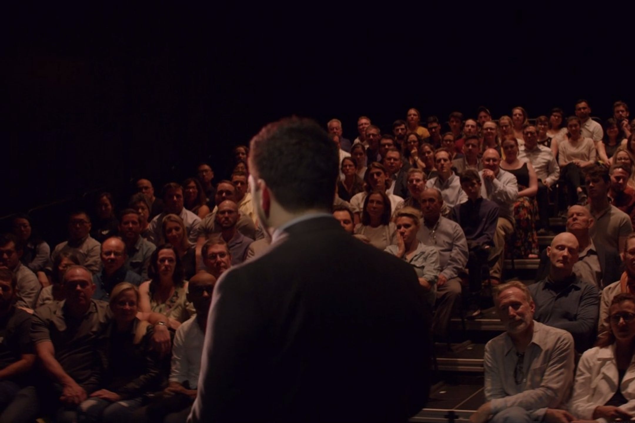 A theatrical audience, viewed from the stage. Kate McKinnon and other celebrities are visible in the crowd.