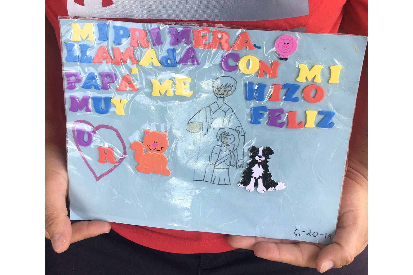 Hands hold up a laminated piece of paper featuring block letters, stickers, and a drawing.