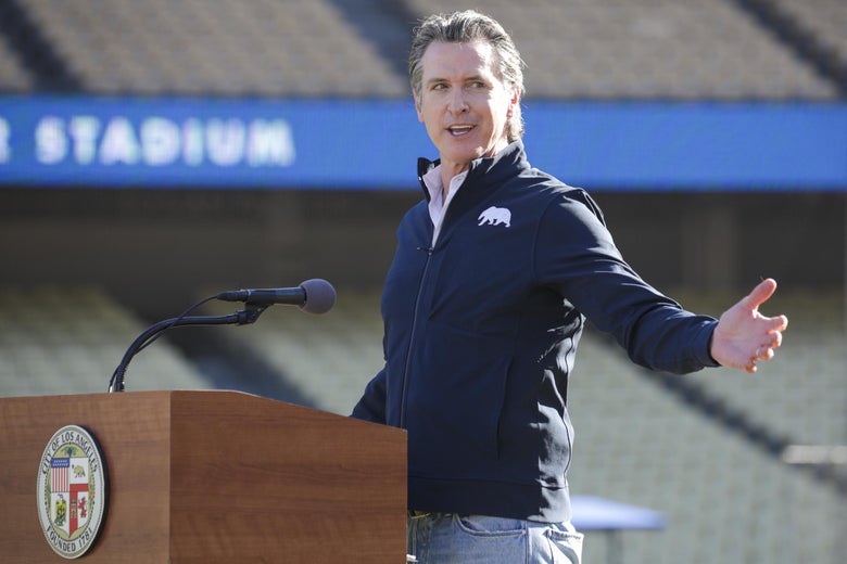 Gavin Newsom stretches his arm out as he speaks from behind a podium on the field in an empty baseball stadium.