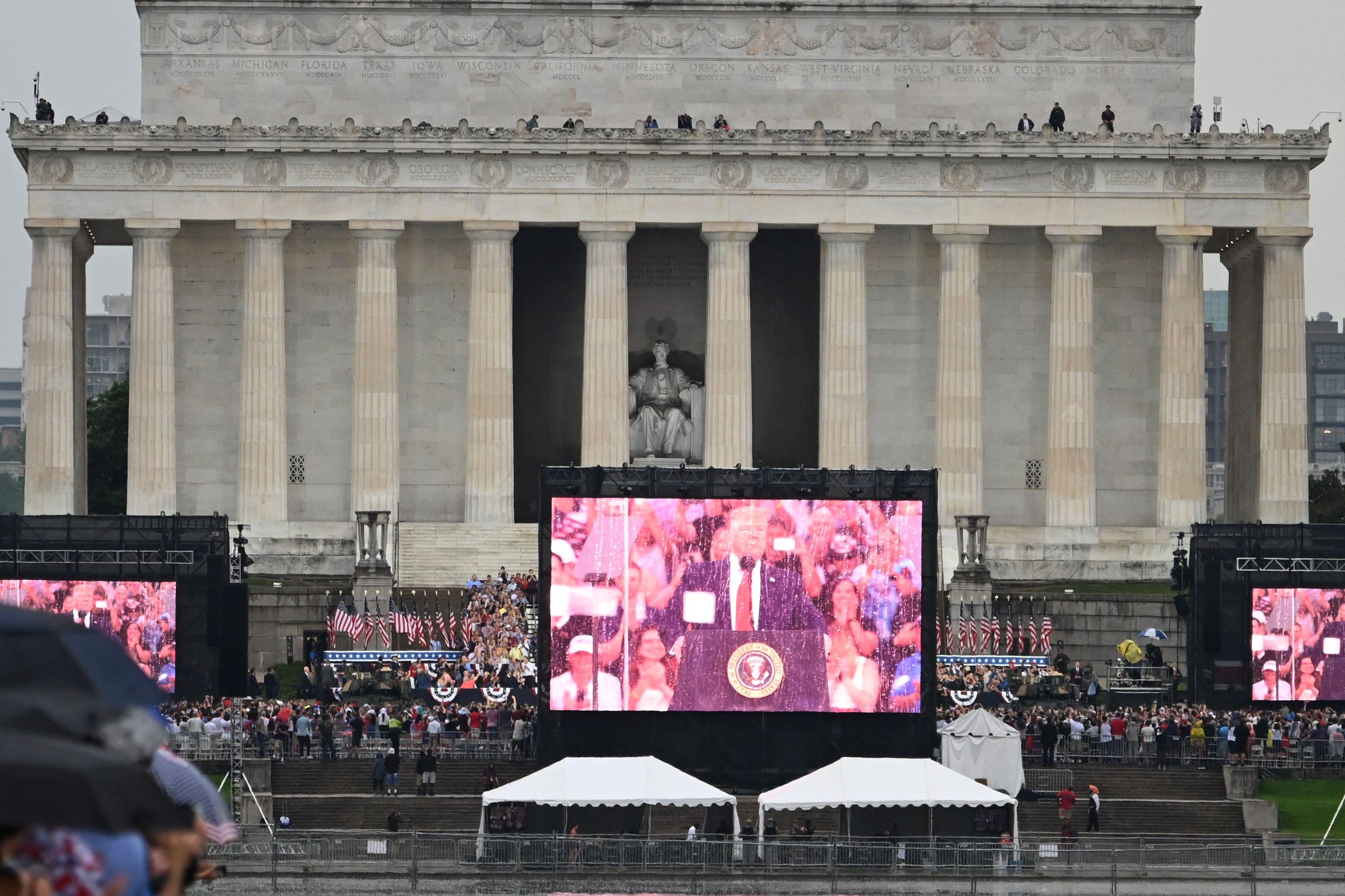 People gather on the National Mall near a screen showing President Donald Trump delivering a speech during the "Salute to America" Fourth of July event at the Lincoln Memorial in Washington, D.C. on July 4, 2019. 