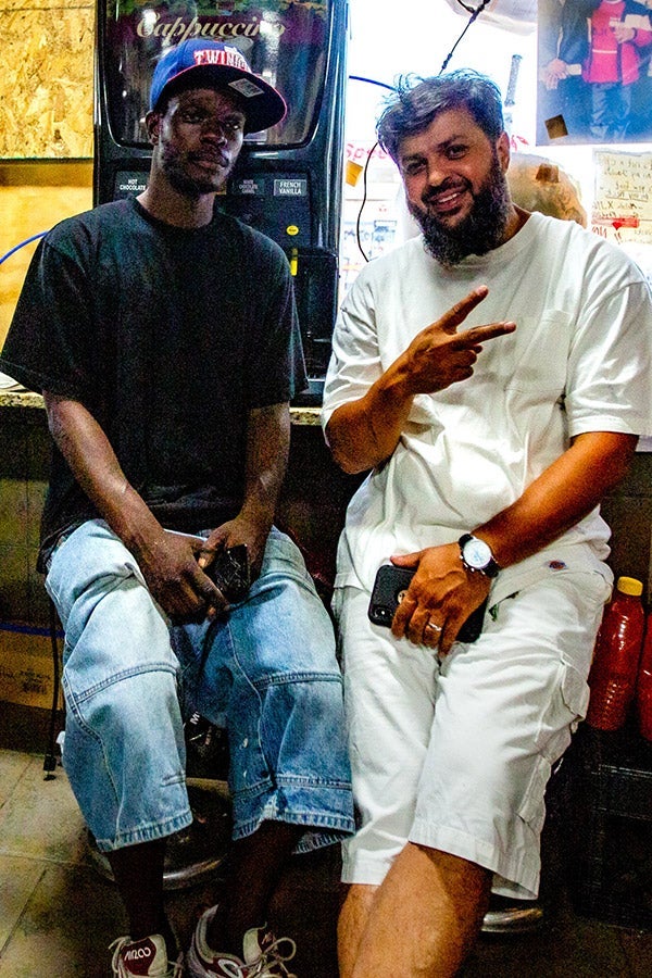 A man in white shorts and a white T-shirt makes a piece sign as a Black man sits beside him.