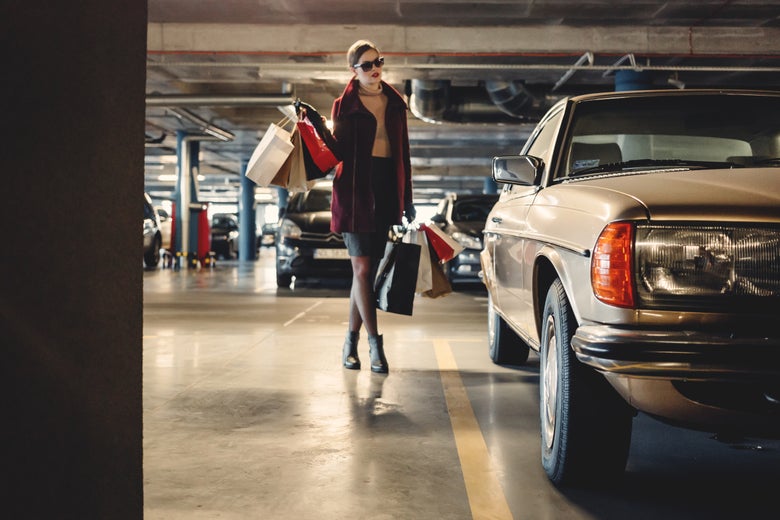 In a parking garage, a fashionable woman carries bags to a car.