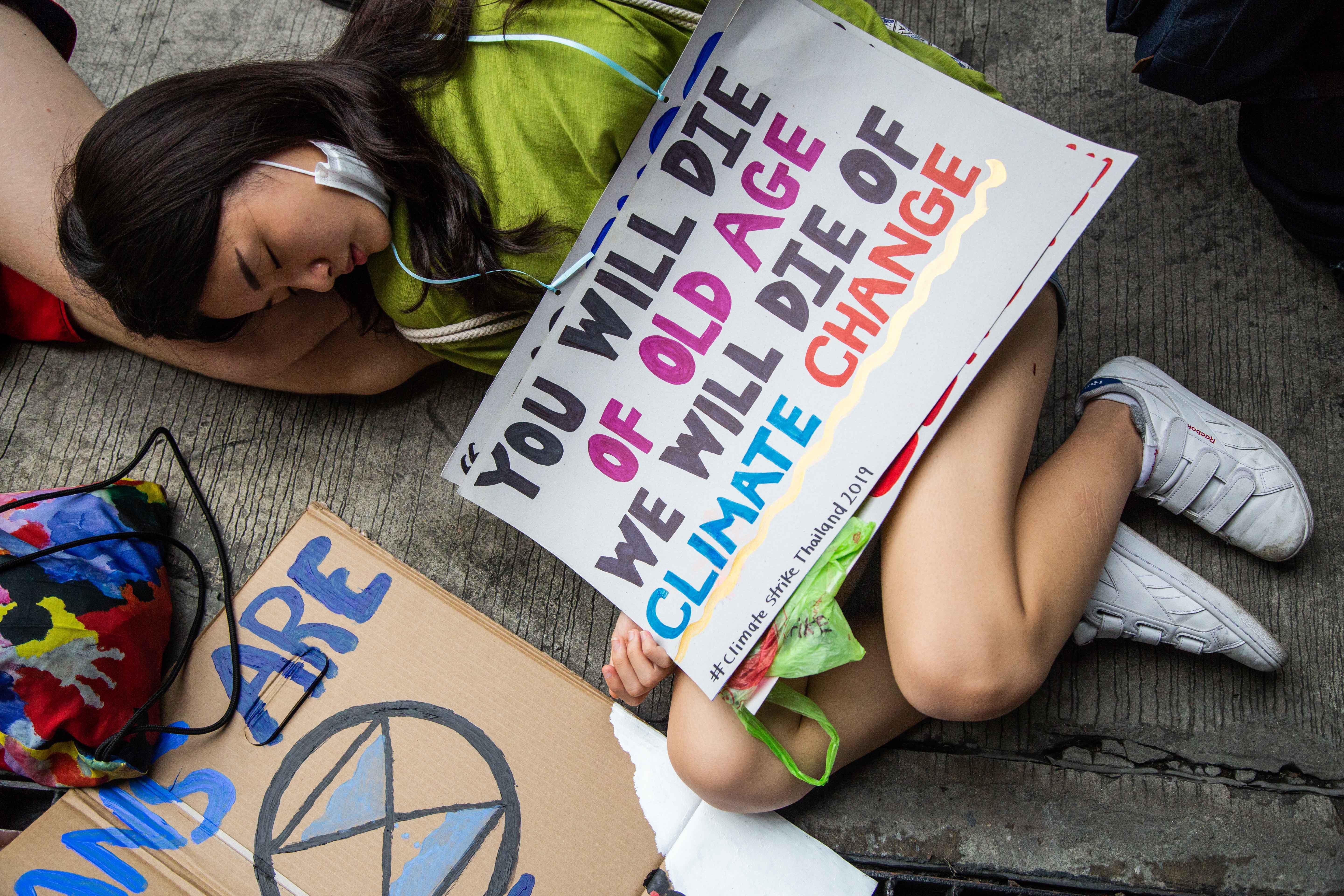 A girl lies on the ground, under a sign that says "you will die of old age, we will die of climate change."