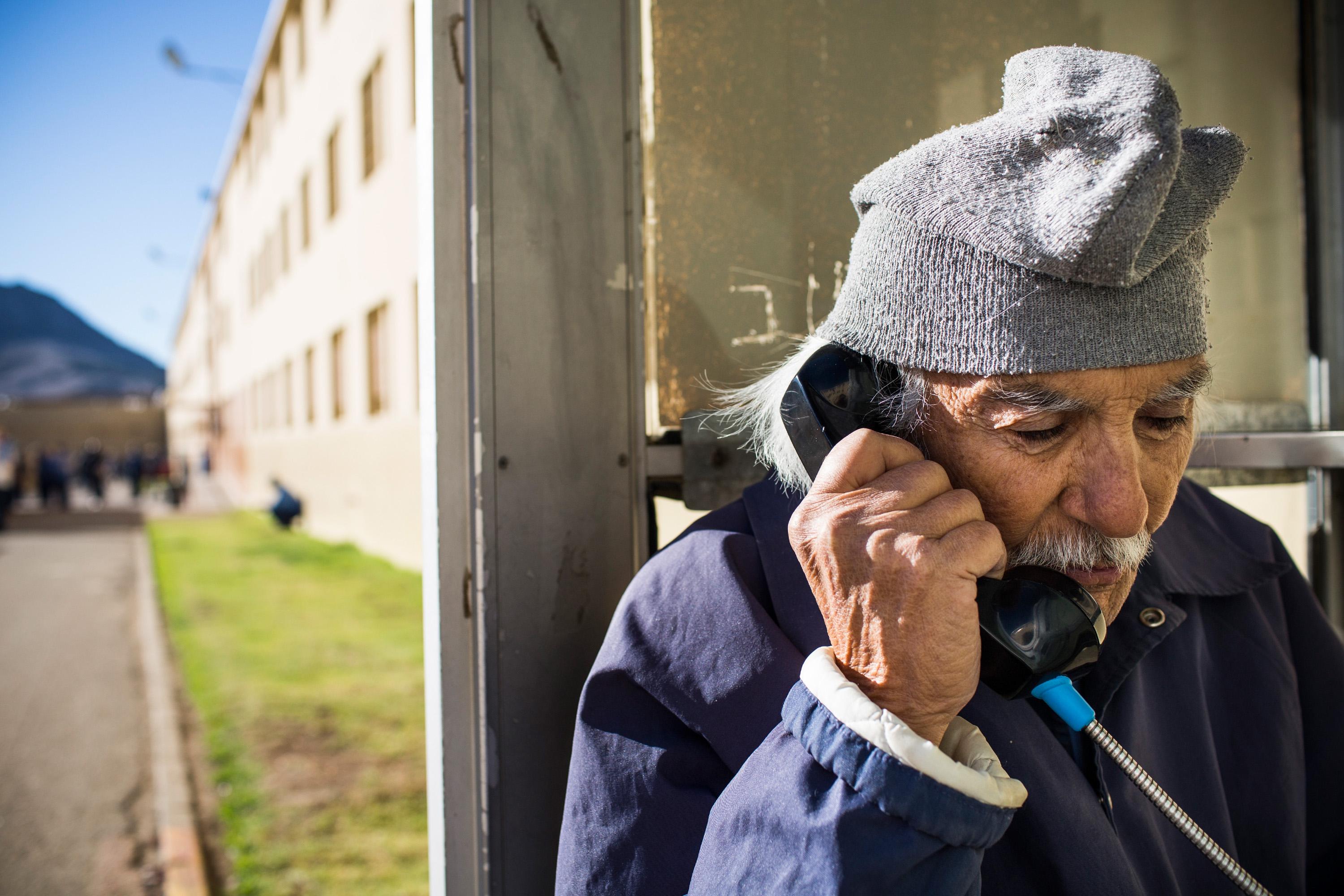An elderly man talks on a wired phone next to a prison yard.