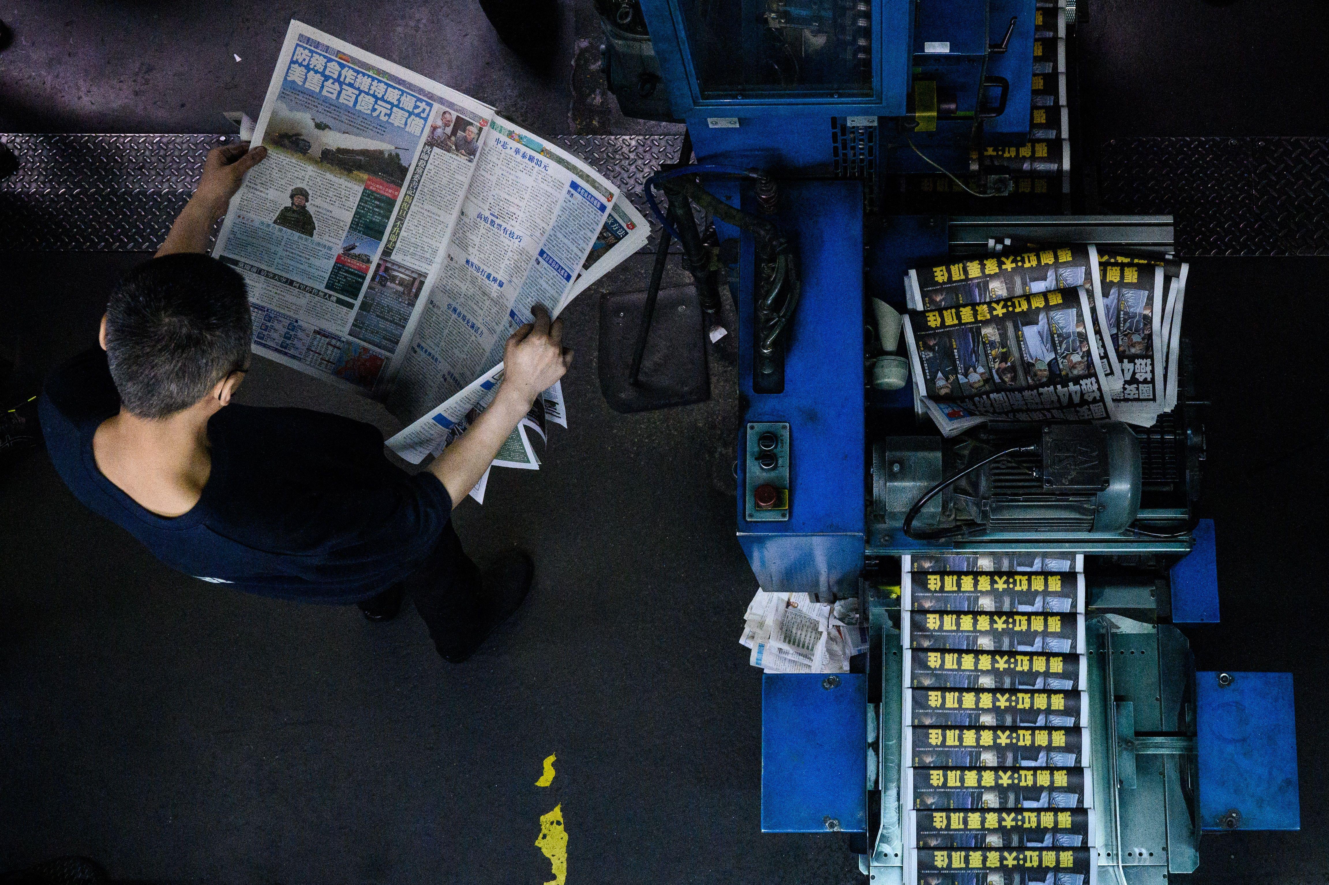An overhead shot of a worker reading the newspaper standing next to the printing press churning out copies of the paper.