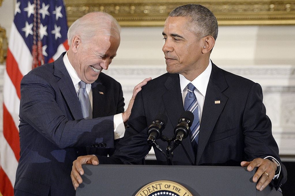 Biden laughs and puts his hand on Obama's shoulder as Obama stands at a lectern.