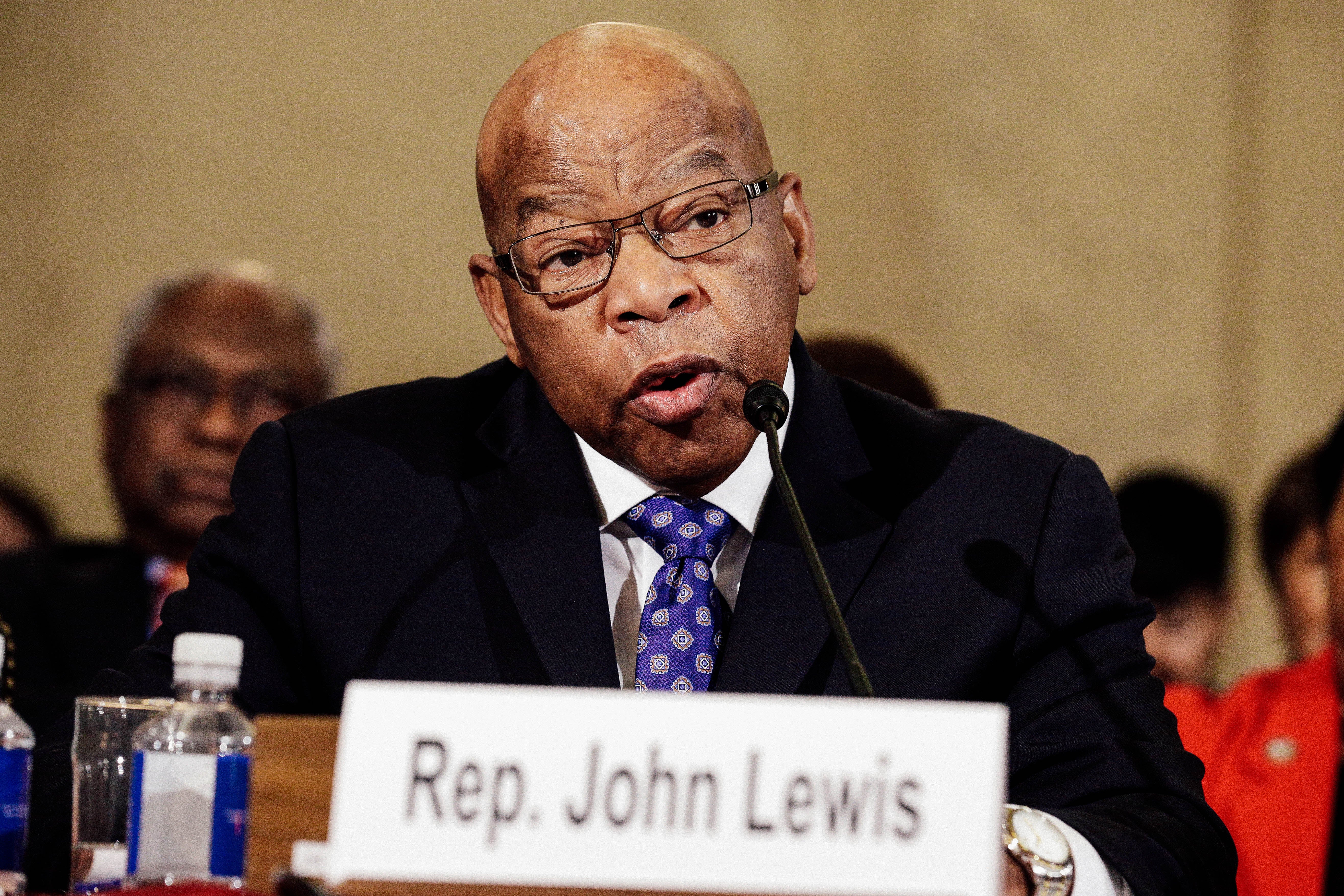 John Lewis speaks at a microphone, with a sign featuring his name displayed in front of him.