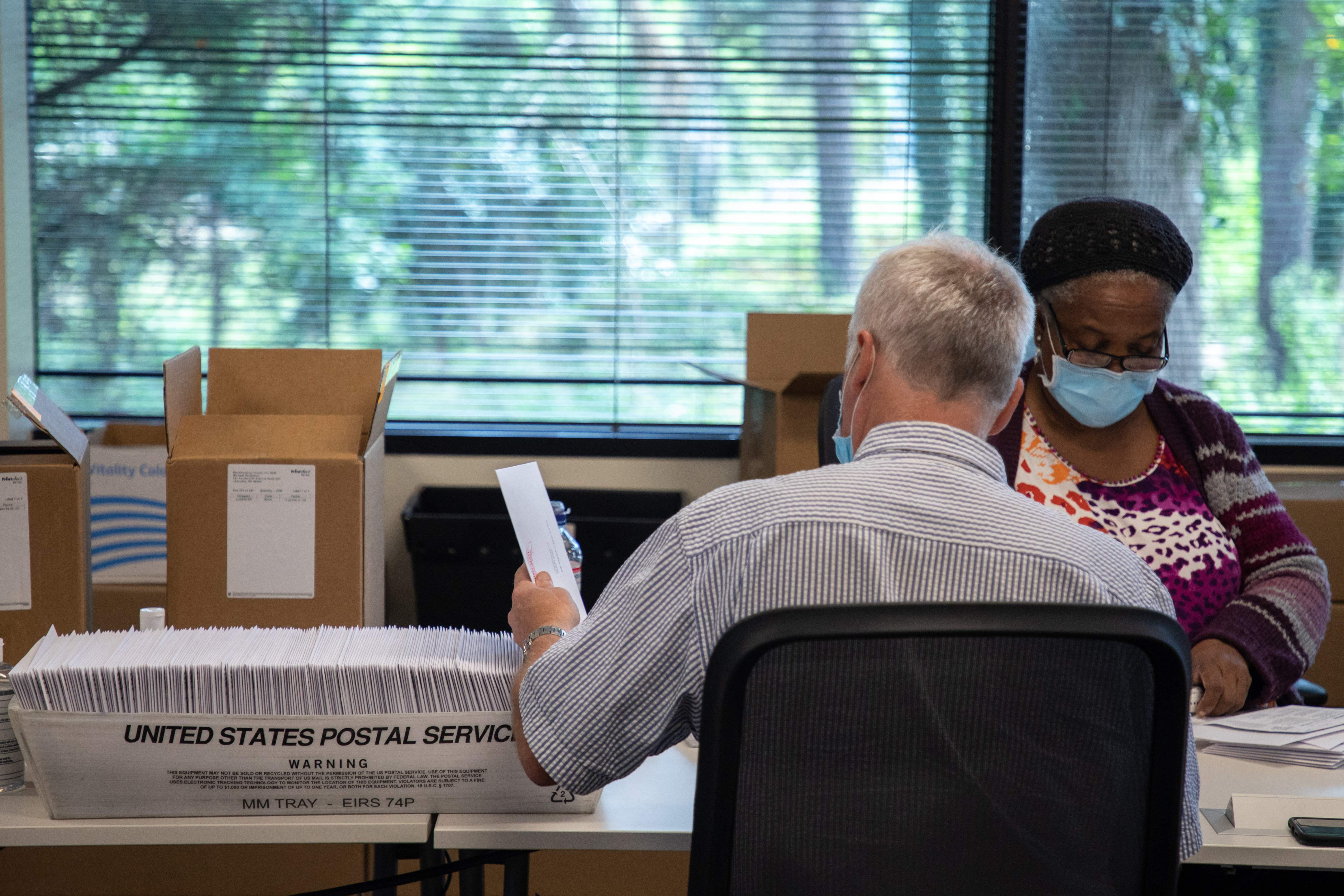 Two election officials sit at a table with boxes of absentee ballot materials and USPS containers.