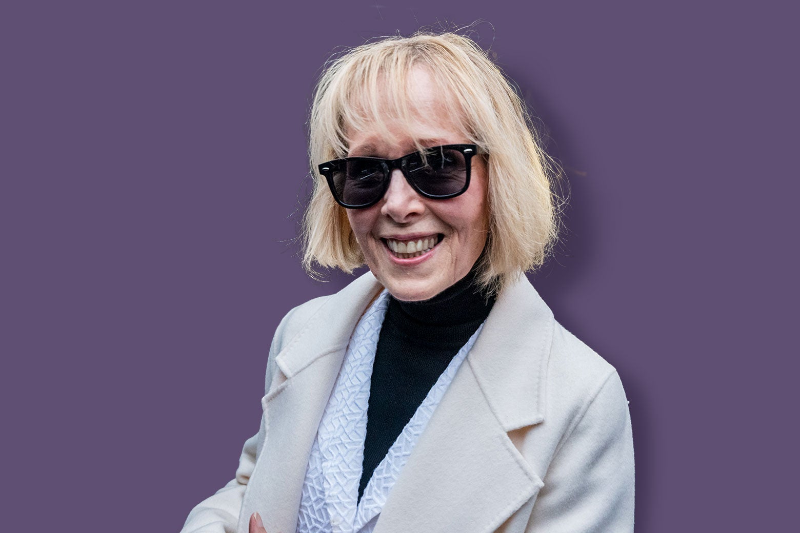 E. Jean Carroll, wearing a black turtleneck and cream coat and dark sunglasses, smiles at the camera in a photo taken after she left the courthouse after her win. 