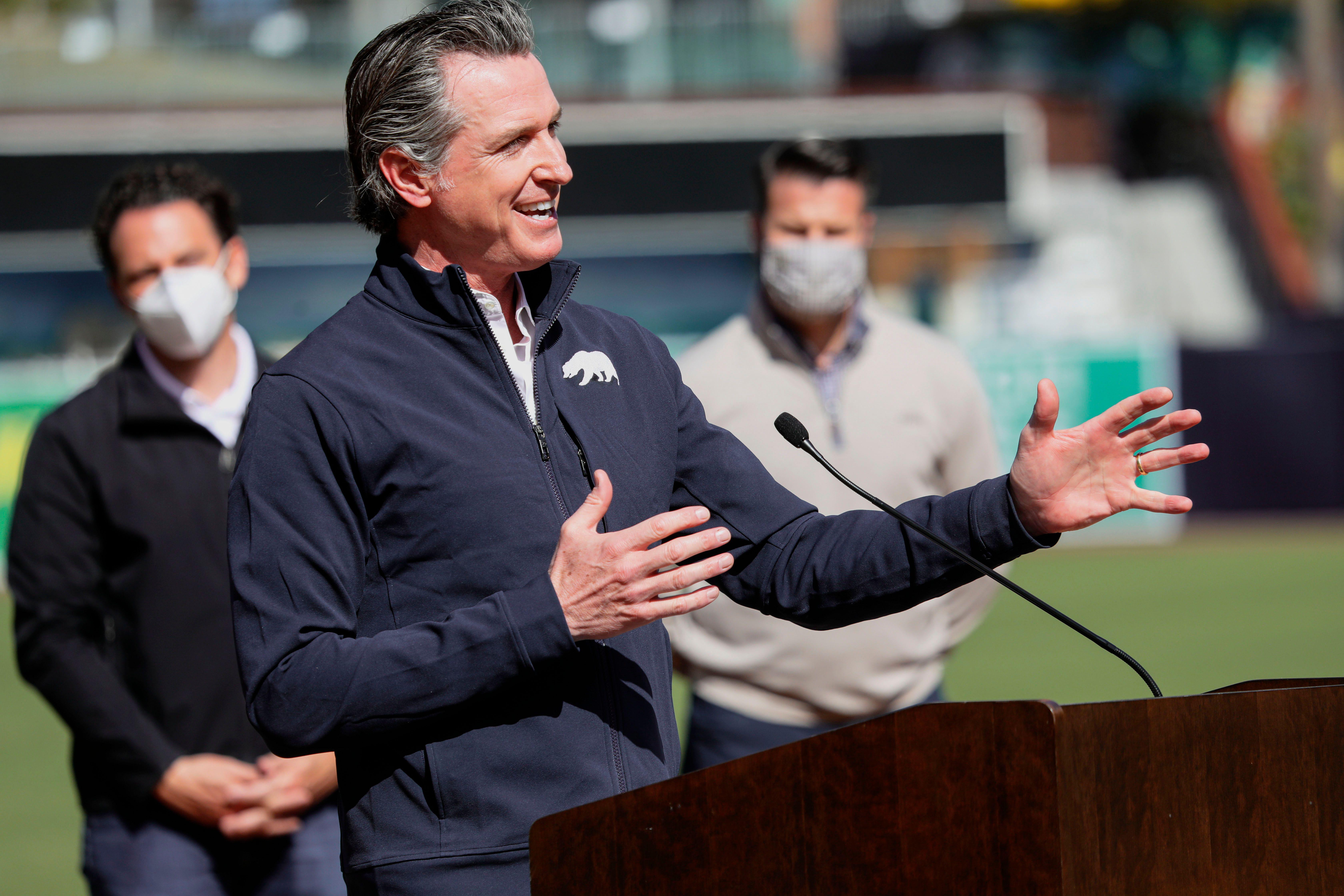 Newsom smiles and gestures as he speaks at a podium on a baseball field, with two men wearing masks in the background