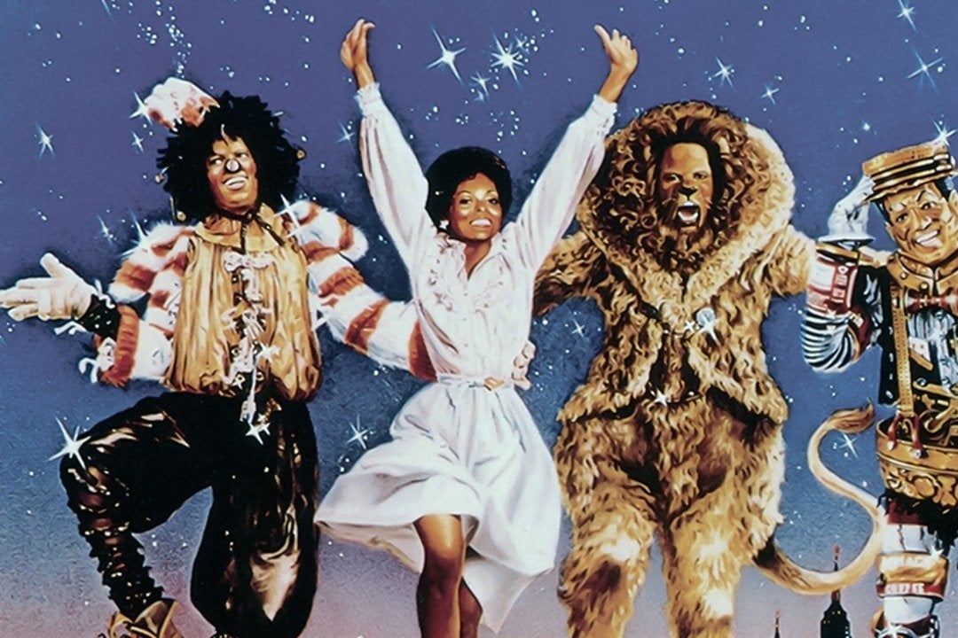 Four people jump up against a blue sky background. One wears a scarecroew outfit and a black wig; another wears a white dress; another is a humanoid lion; another is a robotic fellow. 