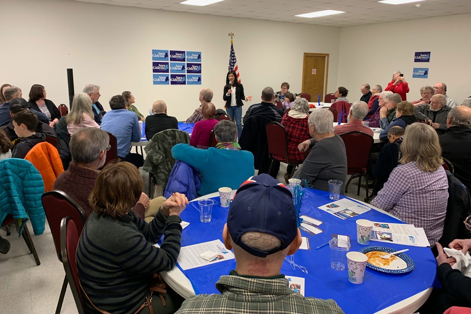 Sara Gideon stands in front of a room with people seated around blue round tables.