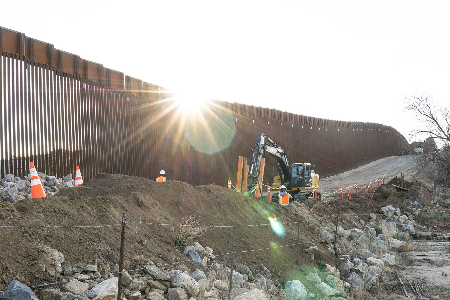 Late sun flares through the top of the United States border wall as a crew works around an excavator, in a strip of dirt, rubble, and barbed wire. 