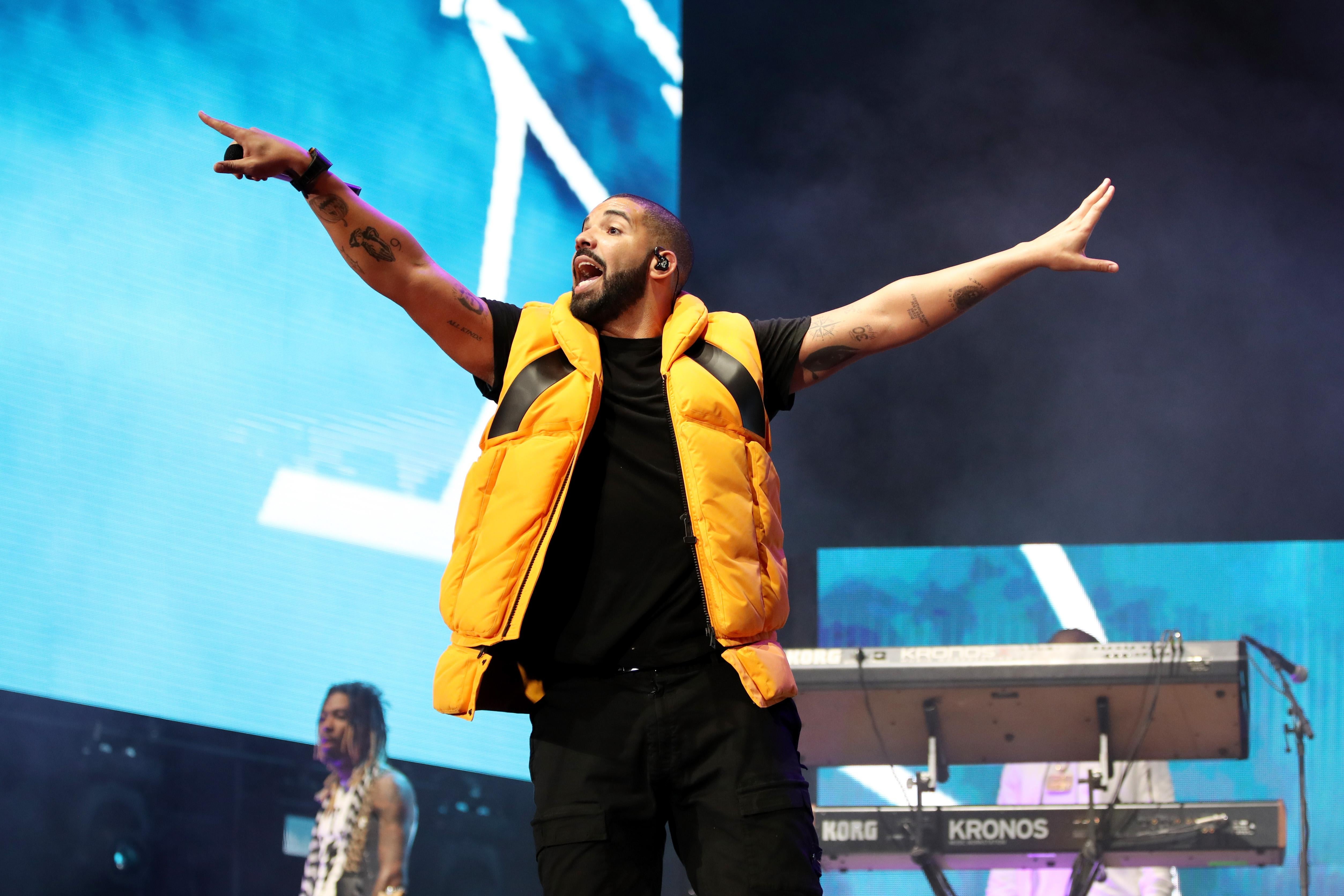 A man in a yellow puffy vest, black shirt, and black pants holds out his arms with his mouth open. He is performing on a stage, pointing a finger out with his right hand toward the audience. 