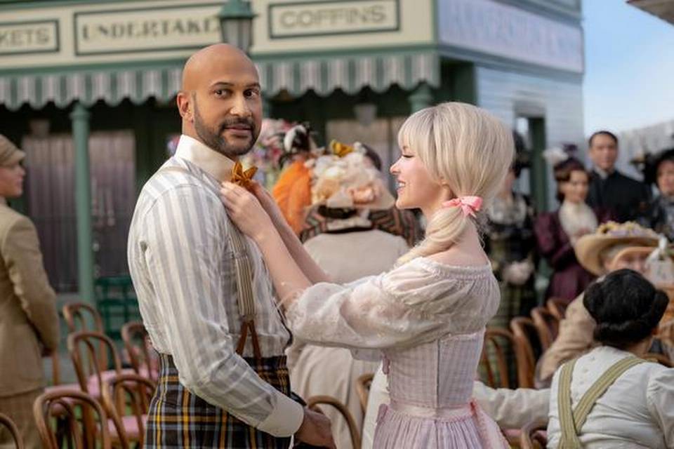 Dove Cameron adjusts the bow tie of Keegan-Michael Key. They are wearing turn-of-the-century garb straight out of a Golden Age musical surrounded by people dressed similarly.