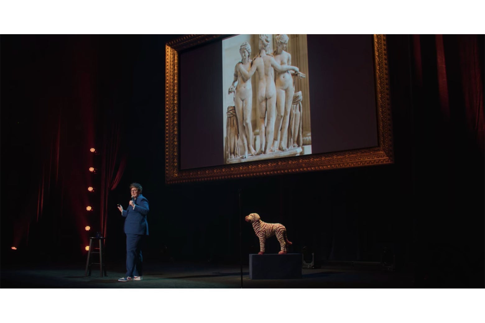 Hannah Gadsby onstage, in front of a screen displaying a photograph of a sculpture of the Three Graces.
