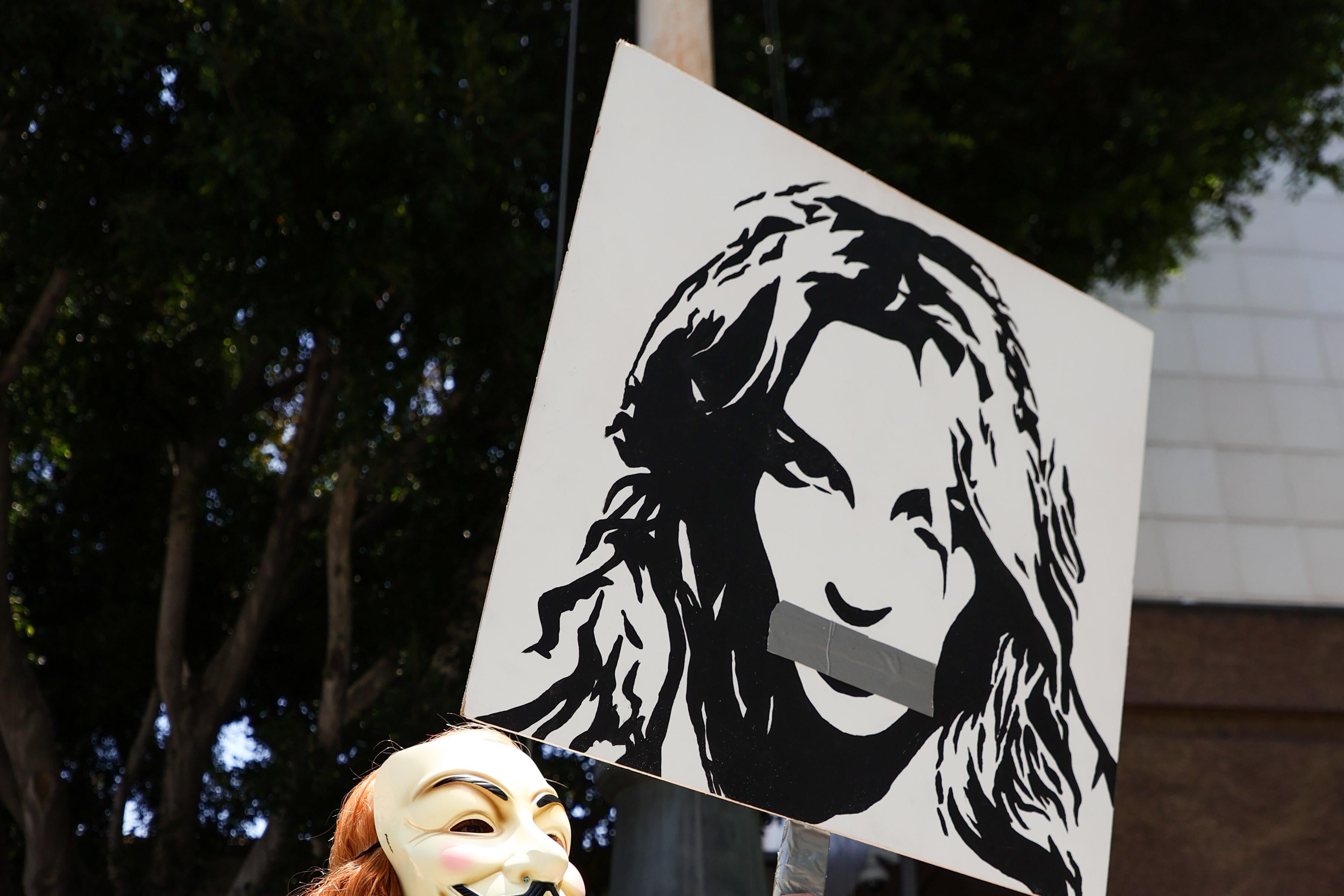 Person with red curly hair wearing a Guy Fawkes mask and a red shiny unitard holds a mic in one hand and a poster of Britney Spears with duct tape over her mouth in her other hand