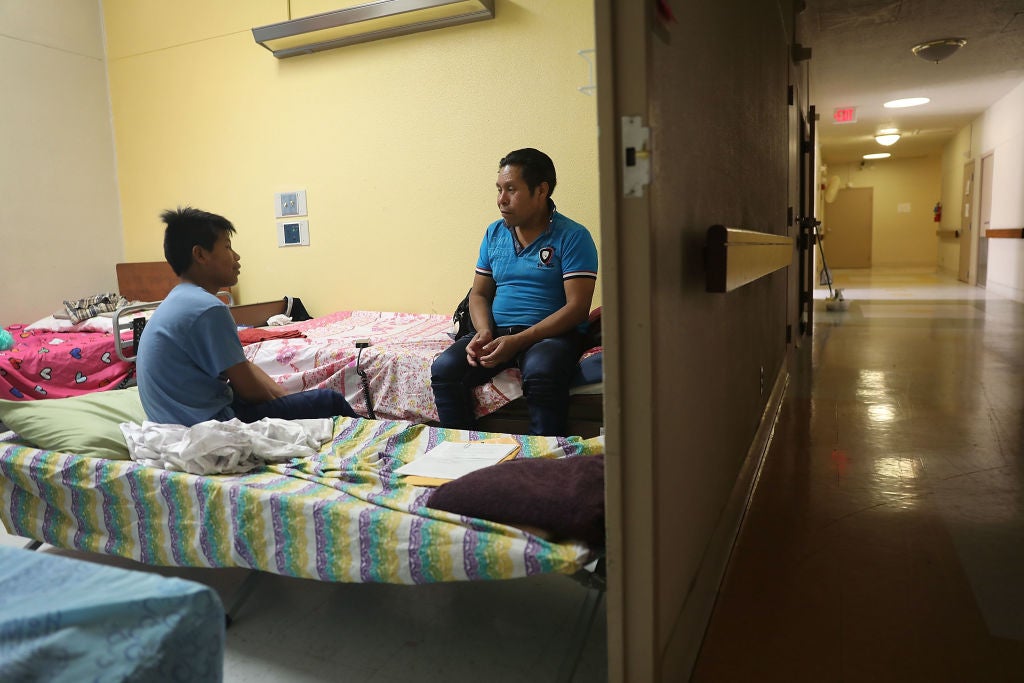 A man and his son speaking to each other while seated on dorm-style beds in a room off of a tile-floored hallway.