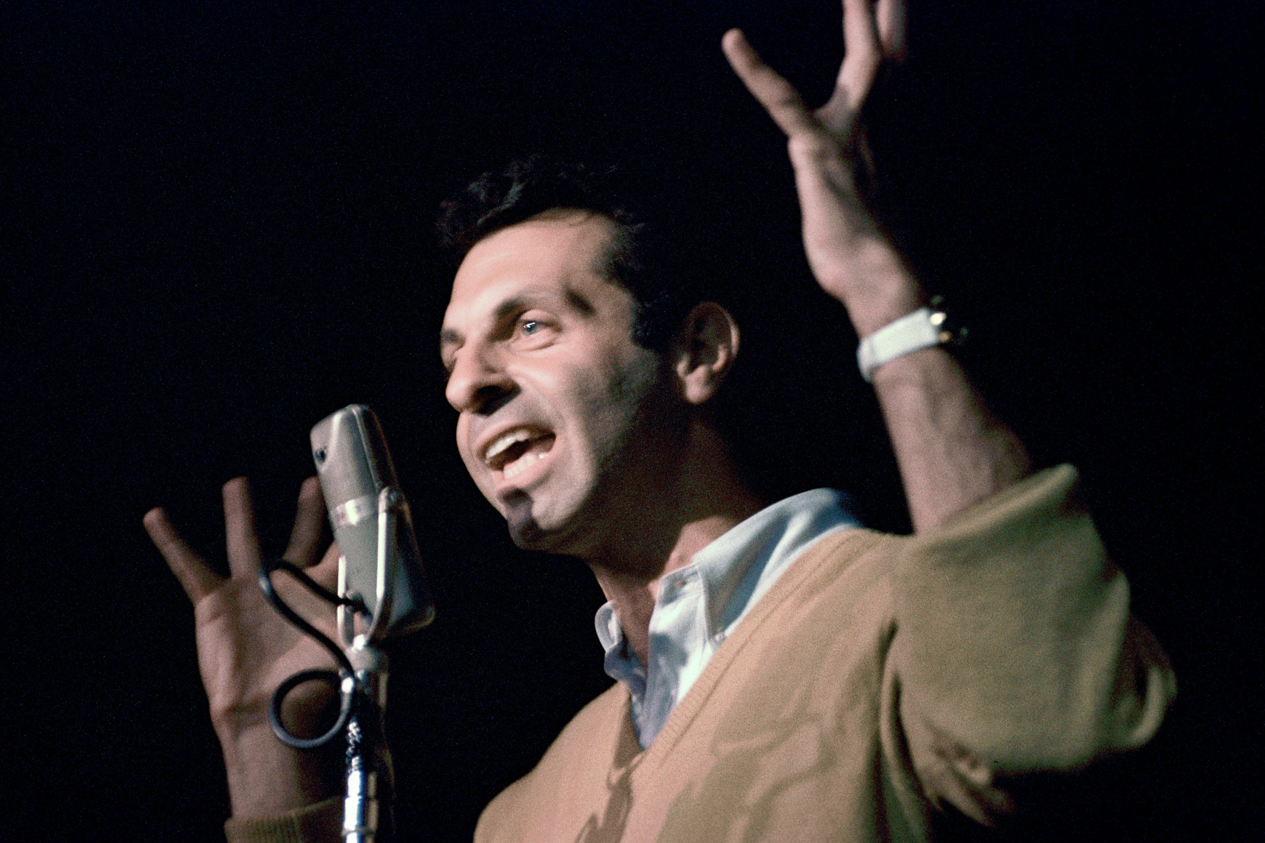A young Mort Sahl speaking into a mic with his hands raised.