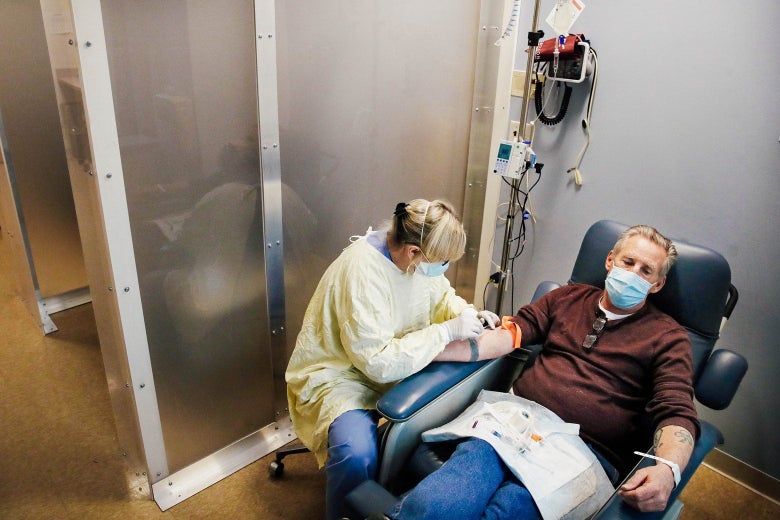 A man wearing a surgical mask sits in a reclining chair receiving an infusion from a nurse in a yellow protective gown