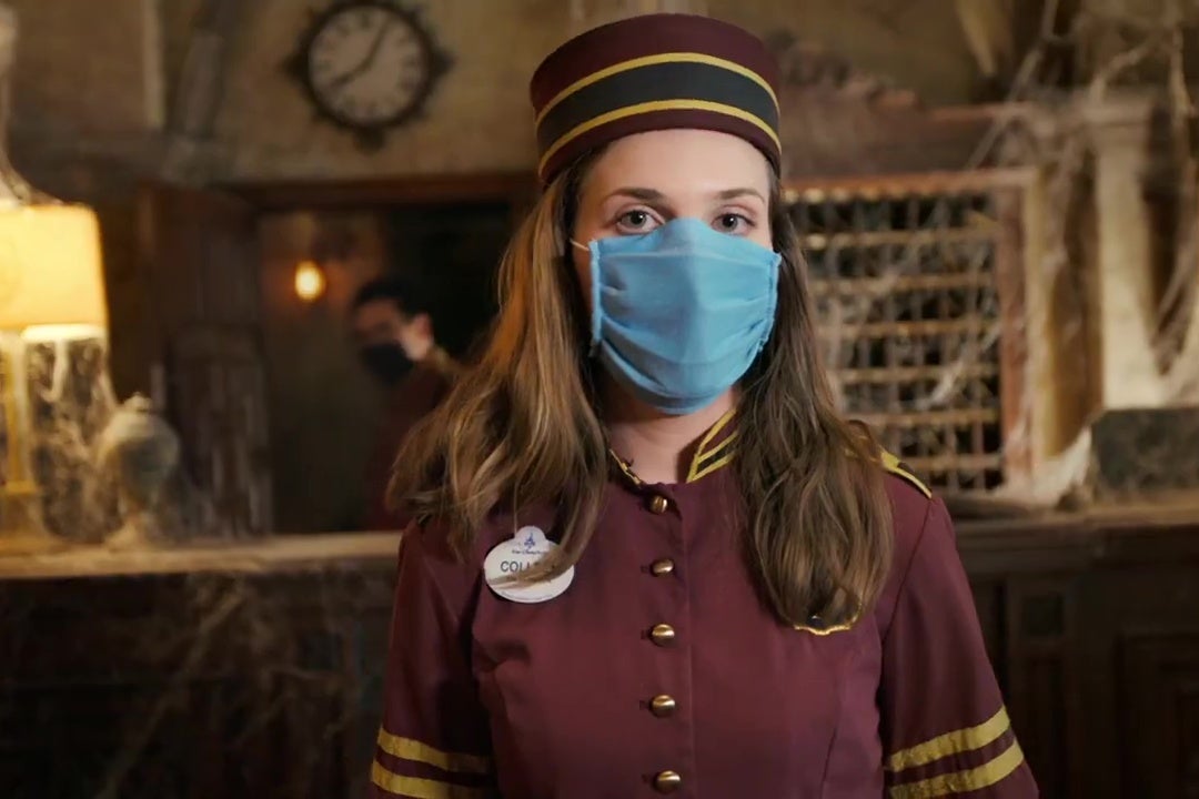 A Disney employee in a 1930s bellboy uniform and surgical mask stands in front of the cobweb-strewn front desk from Walt Disney World's Tower of Terror.