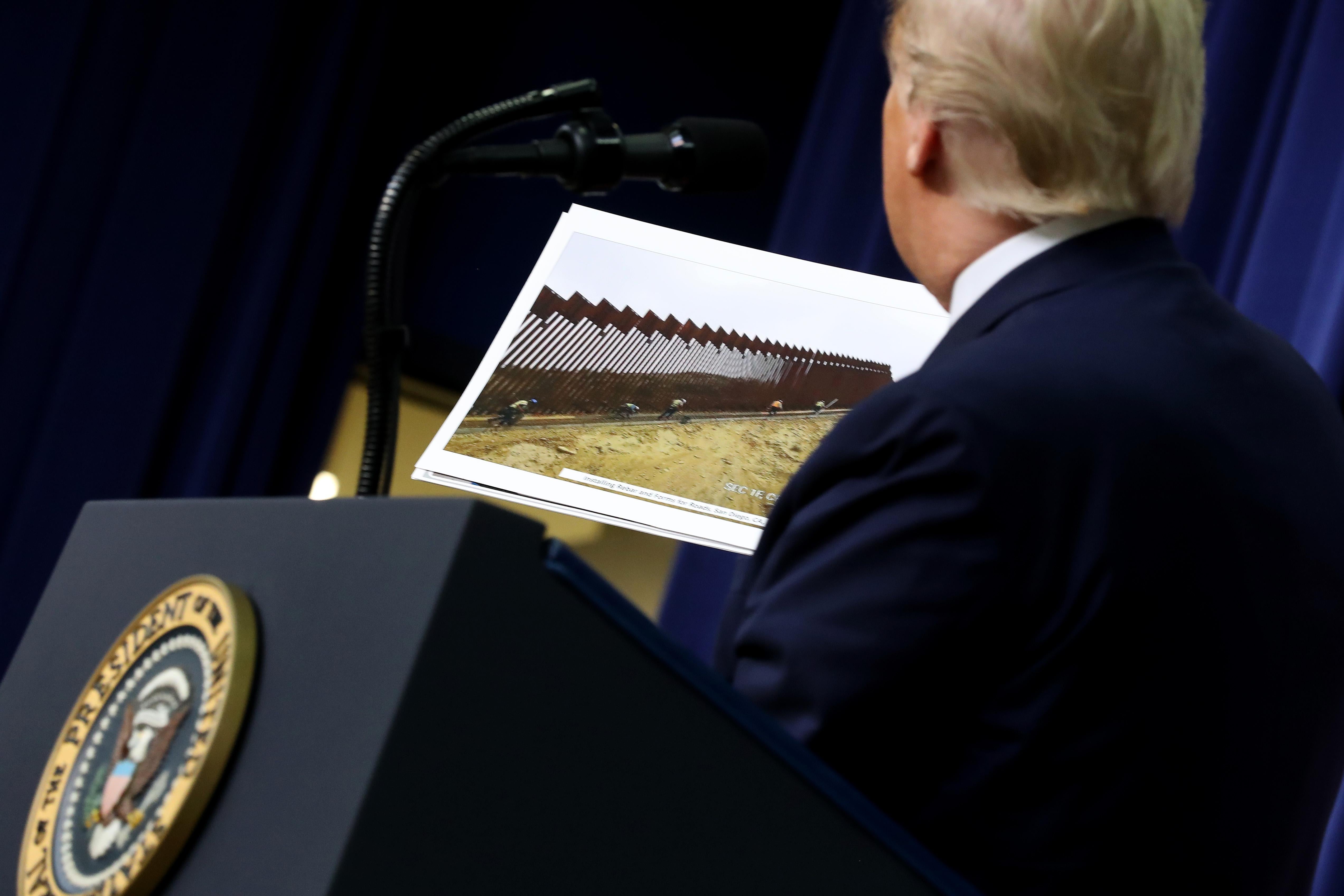 President Trump stands at a podium holding a photograph of border wall construction.