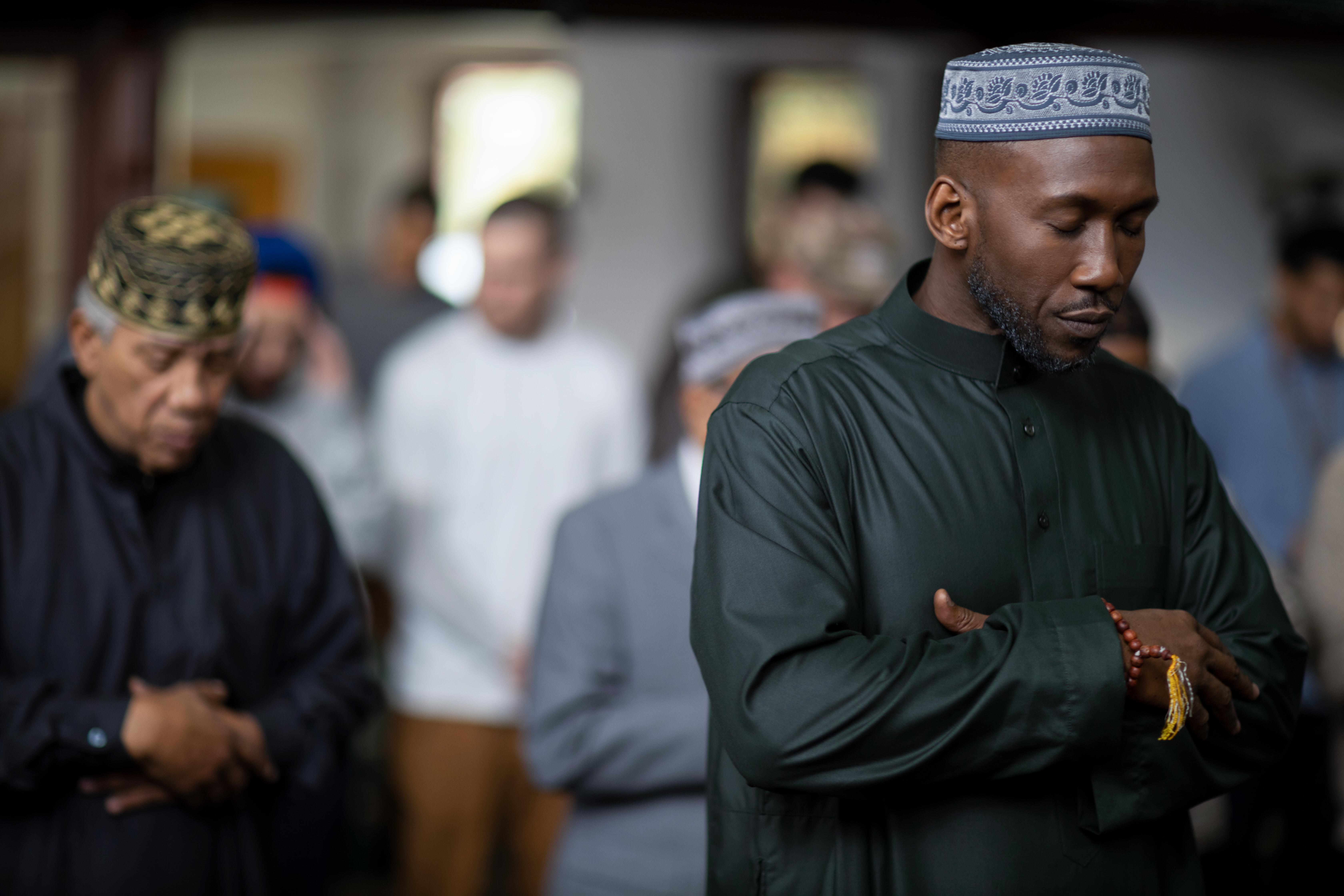 Mahershala Ali folds his hands and closes his eyes. Other Muslims stand behind him.