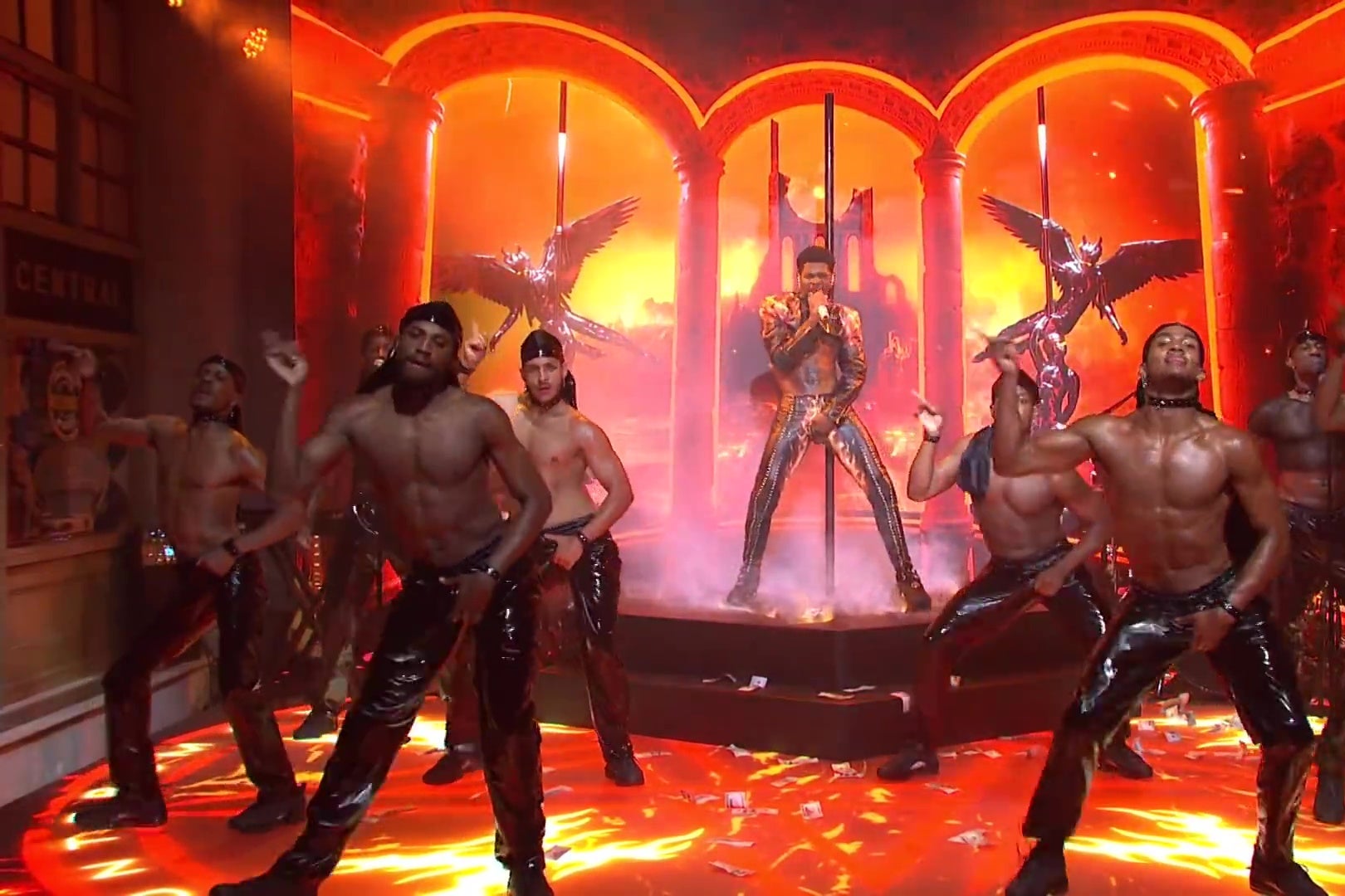 Lil Nas X performs on the SNL stage, which has been made up to look like hell. A line of shirtless black men wearing shiny black pants dance around him.